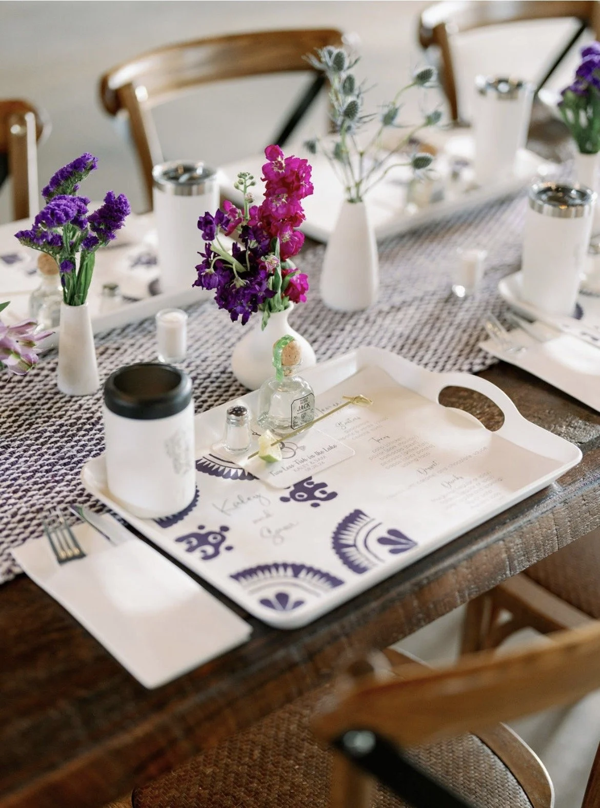 A decorated dining table with purple and pink flowers in vases, white candles, a menu on a tray, and a spray bottle with a cork stopper. The table has a black and white woven table runner, and wooden chairs surround it.