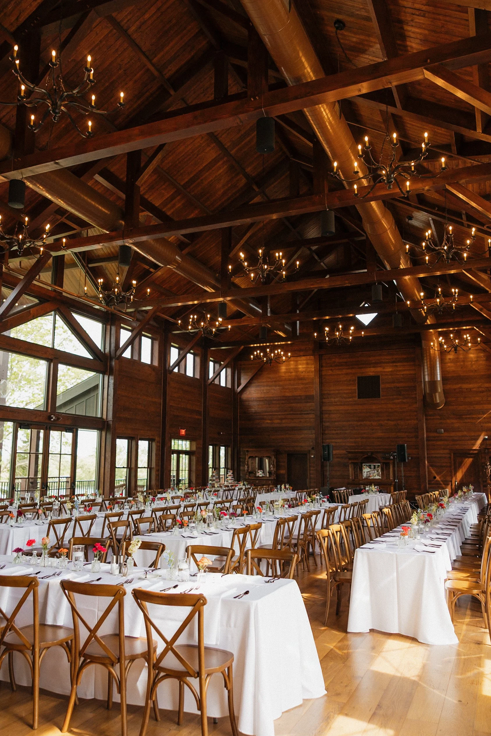 A large banquet hall with a high wooden ceiling and chandeliers, decorated for a formal event with long tables covered with white tablecloths and set with flowers, glasses, and silverware.
