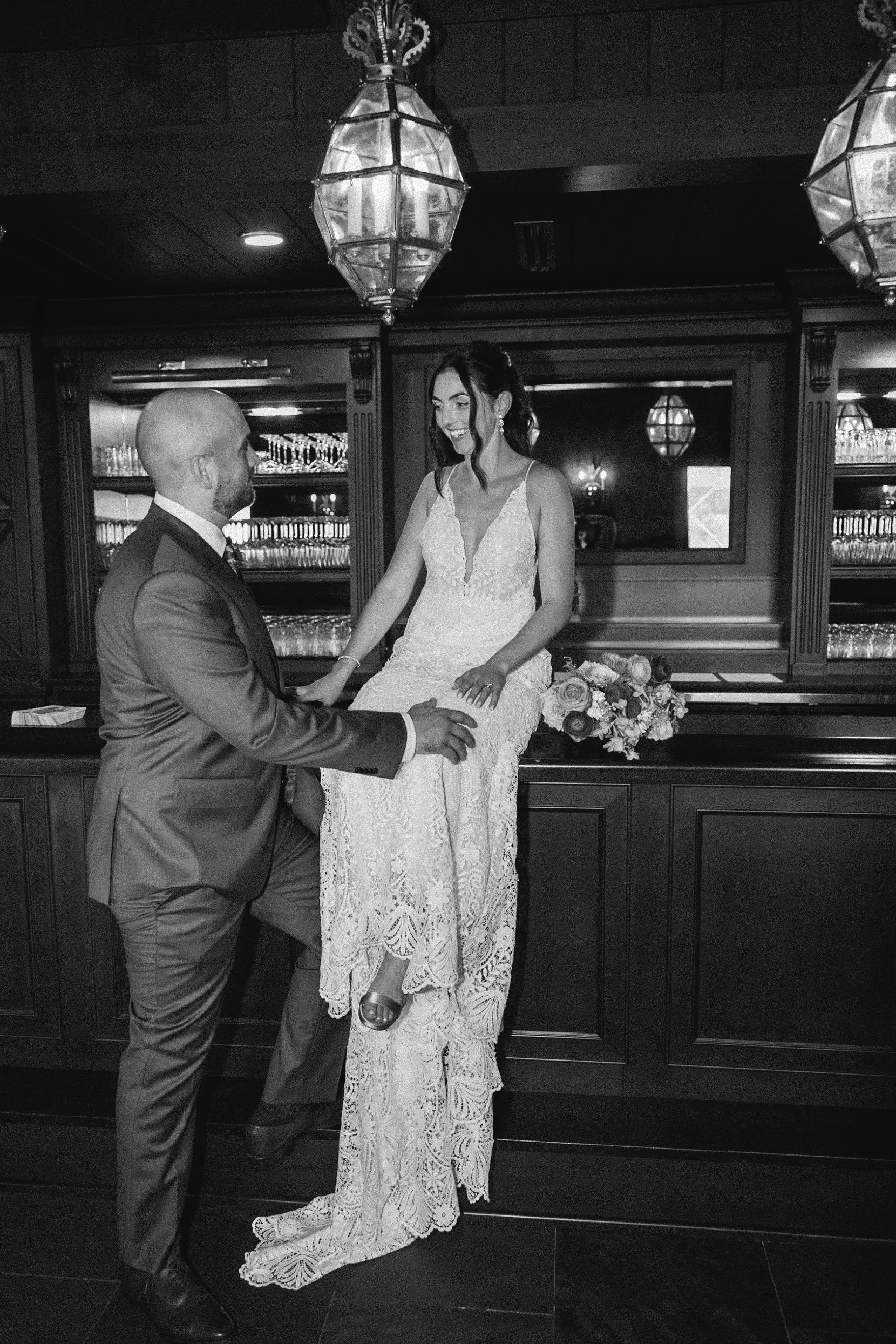 A wedding dress and groom with a tuxedo. The bride is sitting on a wooden surface with a floral bouquet beside her, smiling at the groom who is kneeling in front of her. They are in a room with dark wood paneling and glass shelves behind them.