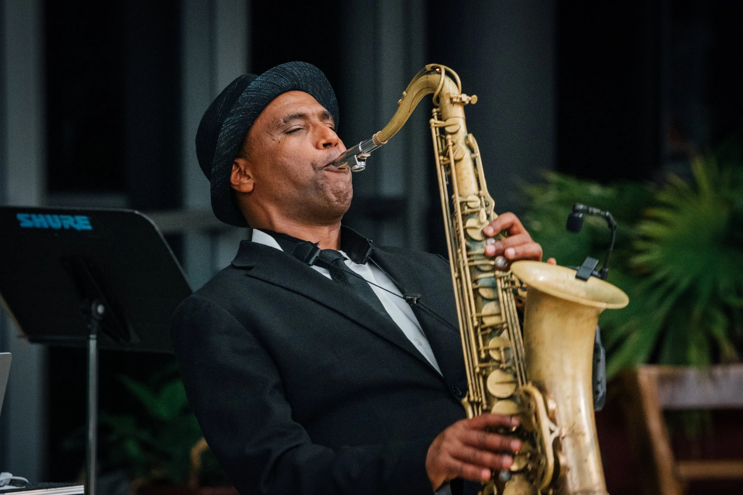 A man in a black suit and hat playing a saxophone.