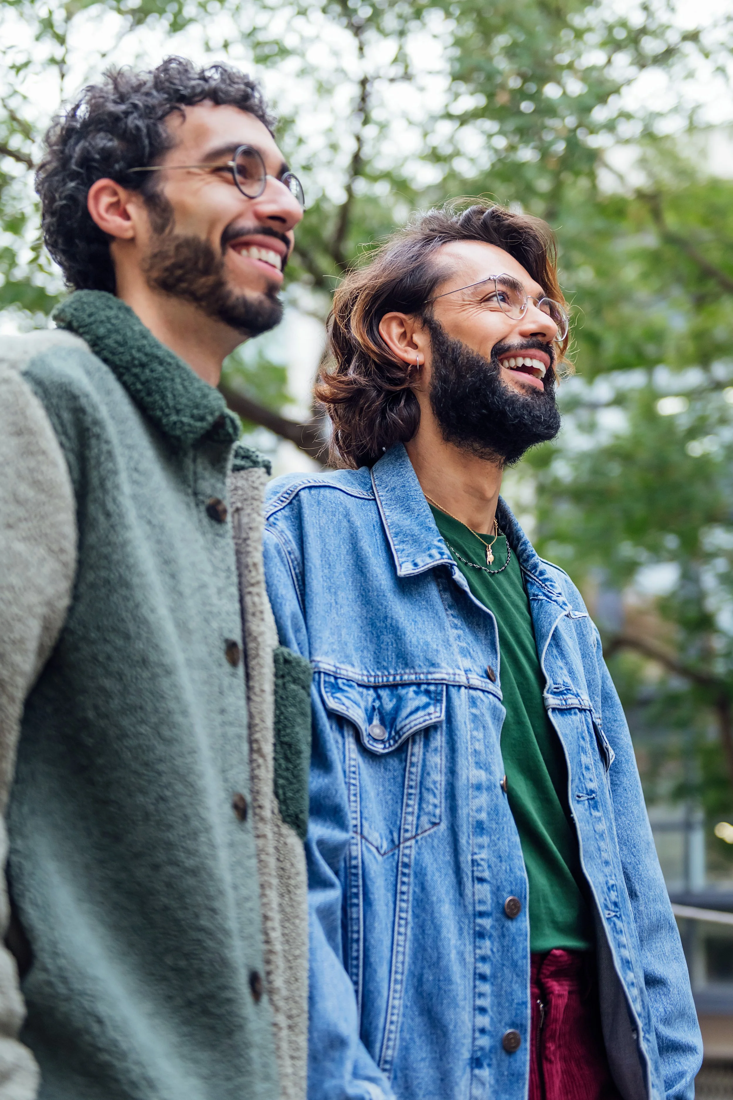 Two smiling men standing outdoors, looking towards the right, with trees in the background.