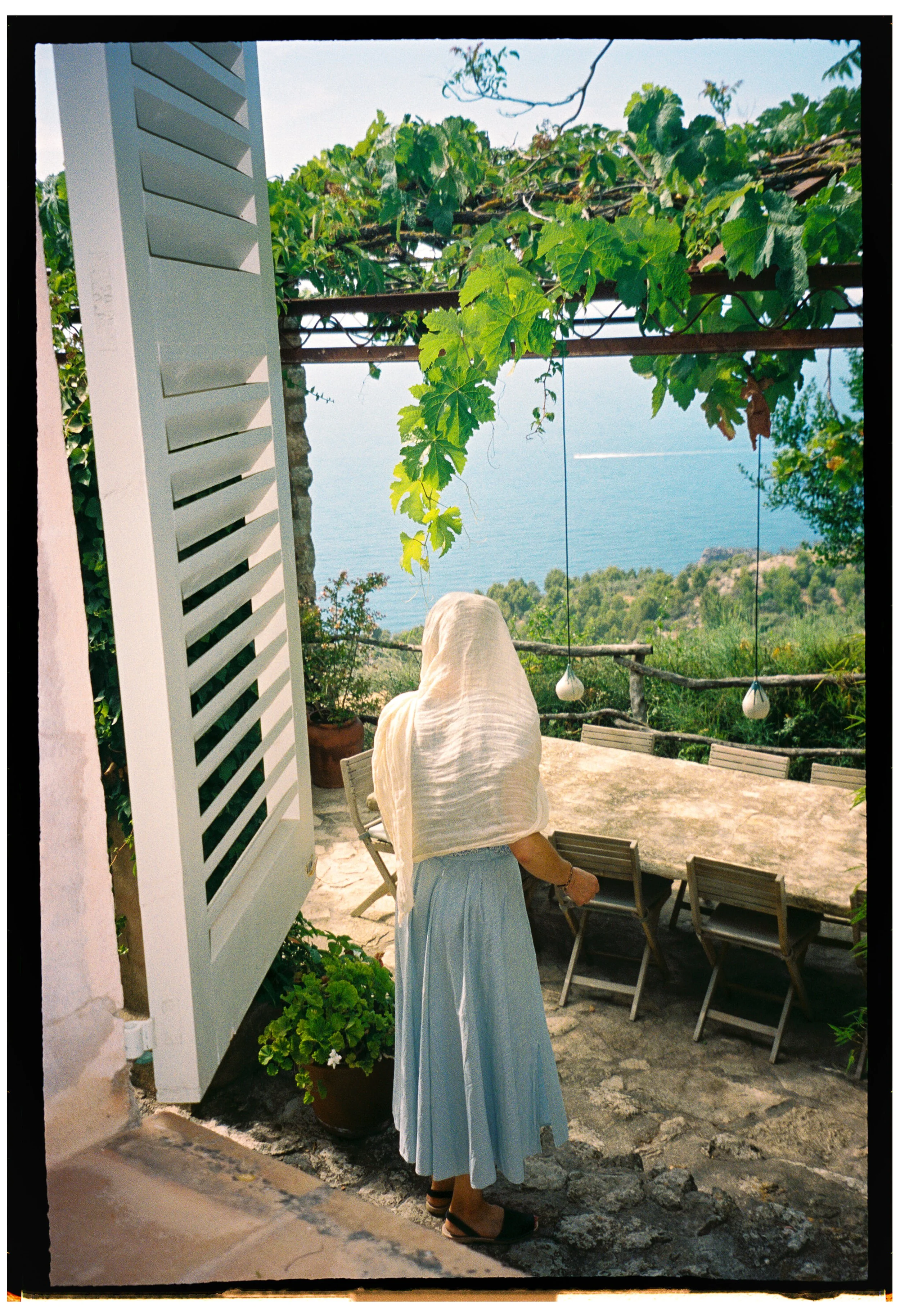 A woman in a long flowing blue skirt and black sandals, with a light-colored scarf, stands on a stone patio overlooking the ocean. She is facing away from the camera and is near a white louvered shutter and potted green plants. In the background, the