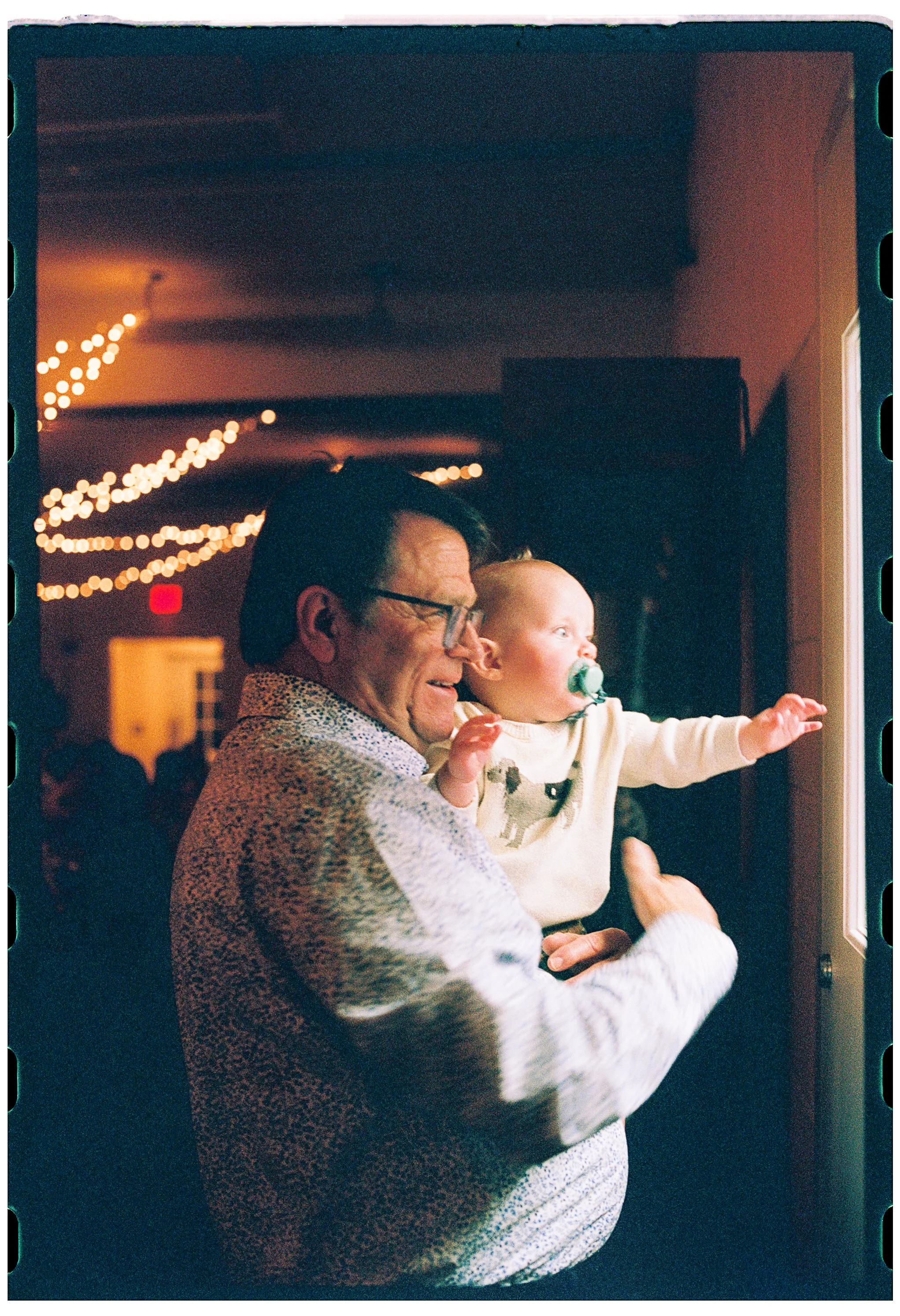 An older woman holding a baby girl while looking out a window. The woman is smiling and the baby has a pacifier. The background has string lights and a ceiling fan.