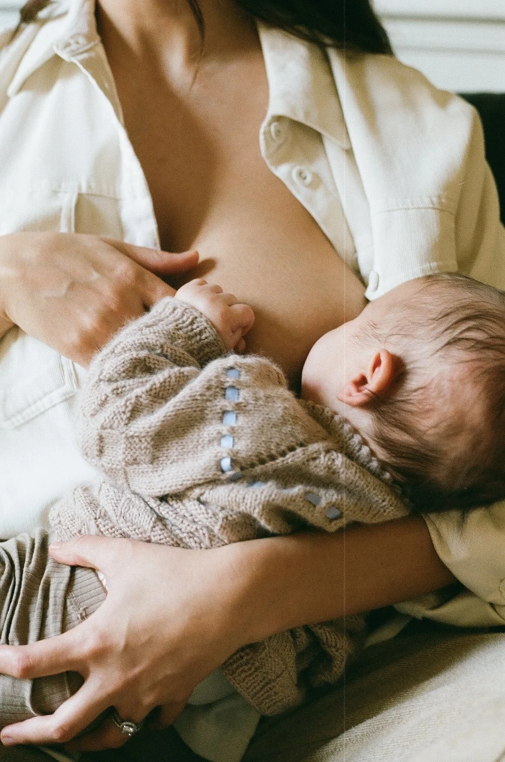 A woman is breastfeeding an infant while sitting down, with her shirt partially open. The baby is wearing a knitted beige sweater with blue accents.