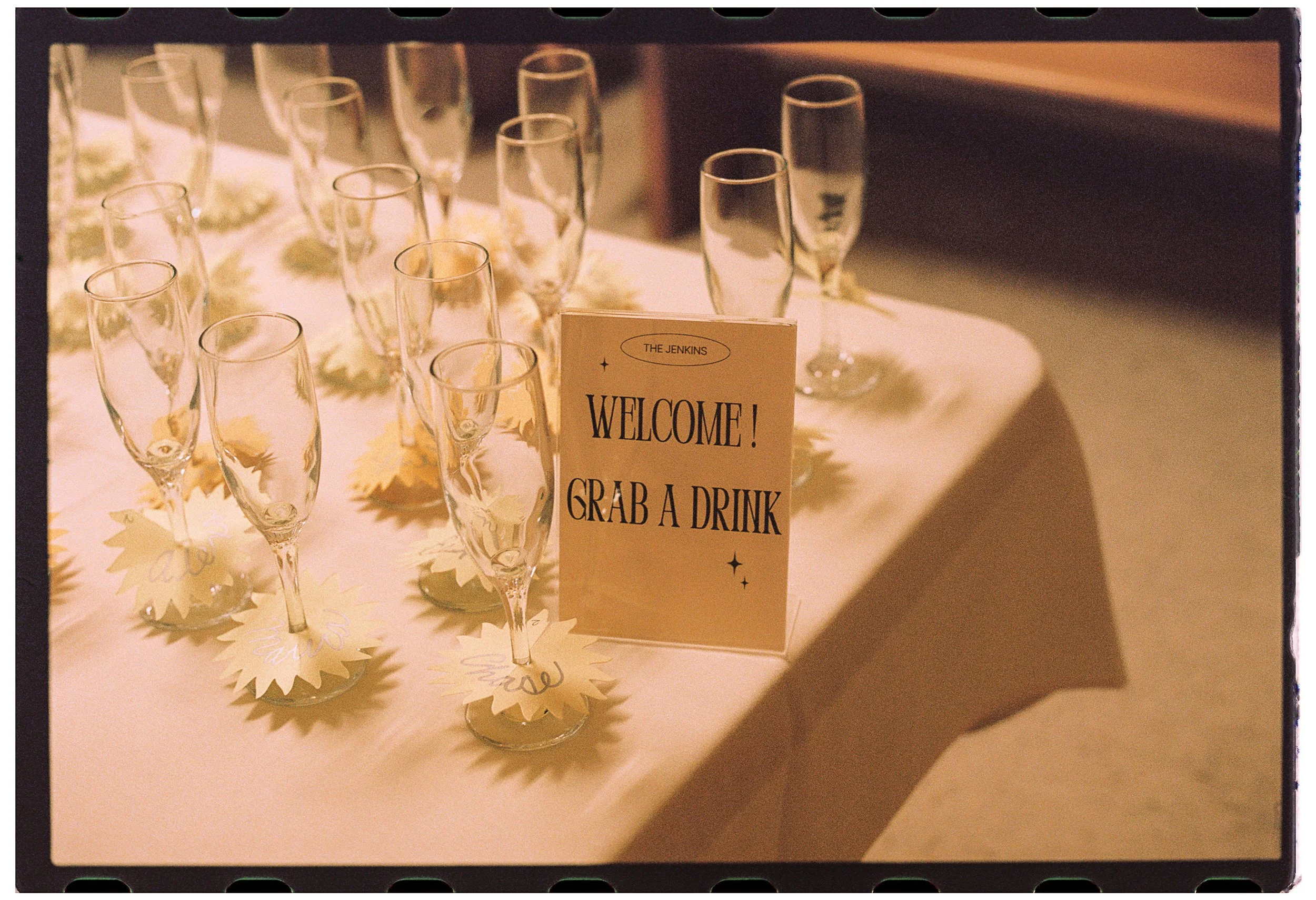 A table set with multiple empty champagne flutes, a framed sign that reads 'Welcome! Grab a drink,' and decorative paper cutouts resembling snowflakes or leaves, likely for a celebration or gathering.