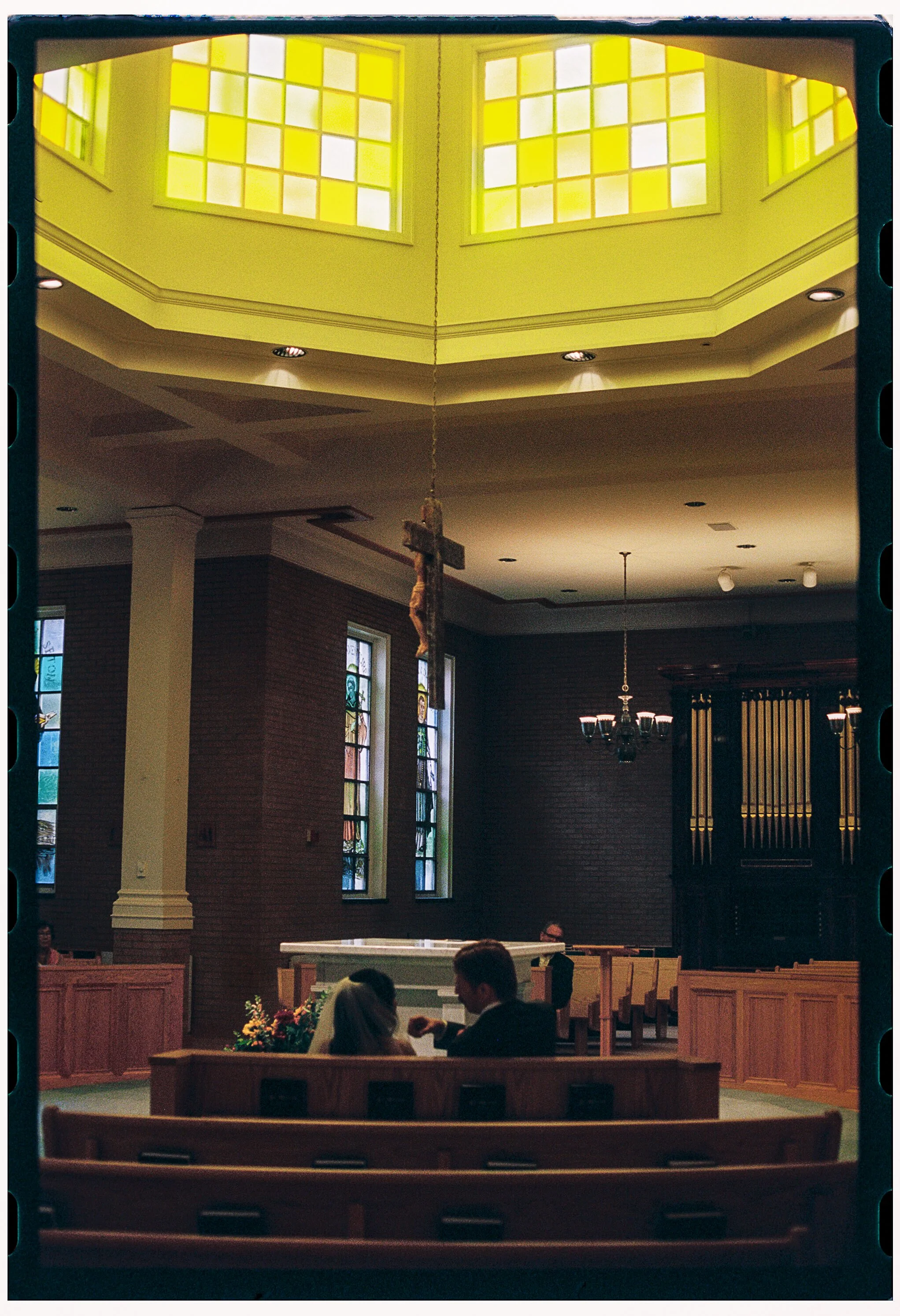 Inside a church or chapel, with stained glass windows, wooden pews, flower arrangements, and a cross hanging from the ceiling. People are seated and engaged in conversation.