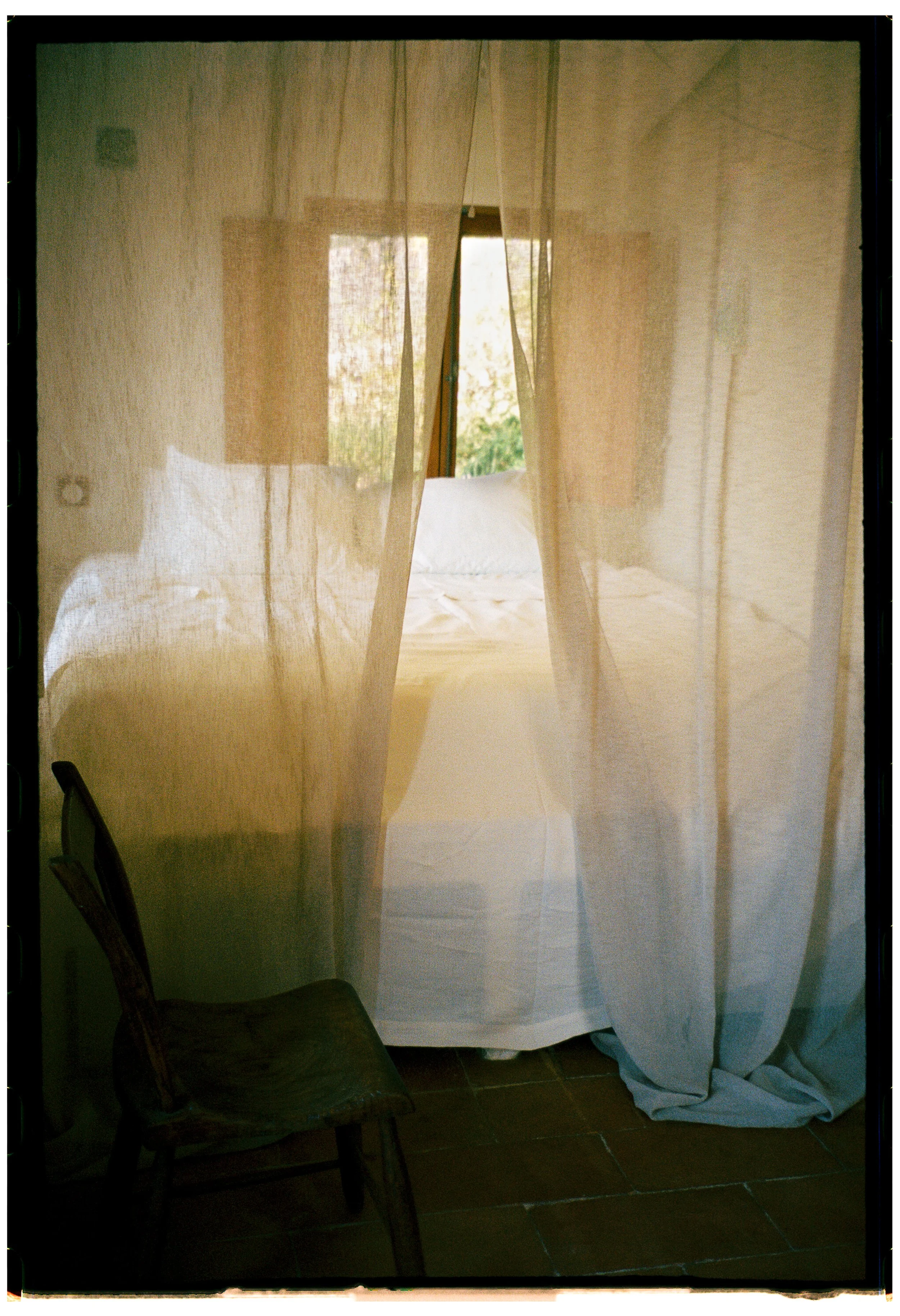 A view through a curtain on a window showing a room with a bed and a window outside, a chair partially visible, and a tiled floor.