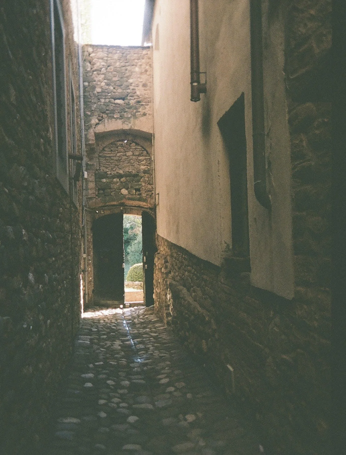 Stone passageway with cobblestone floor and arched doorways, opening to a sunlit outdoor area with greenery.