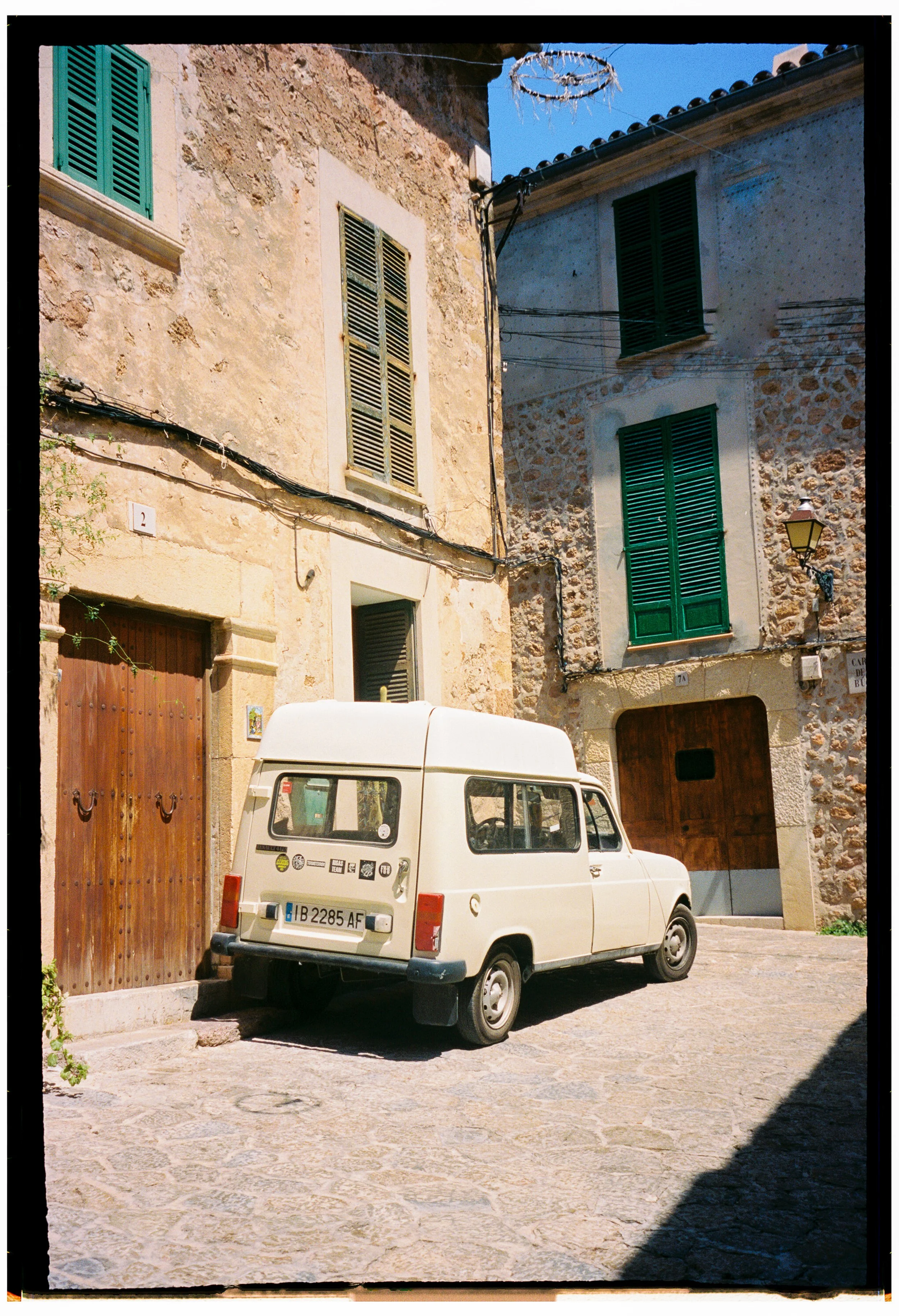 A white vintage van parked on a cobblestone street in front of rustic stone buildings with green shutters and a wooden door.