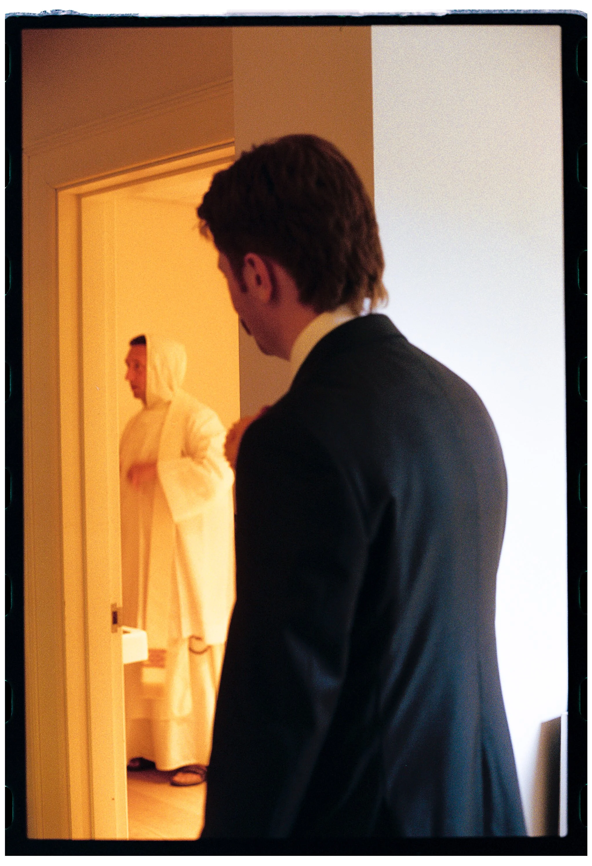 A photo of a man in a suit looking towards a woman in a white robe and hood standing in a doorway.