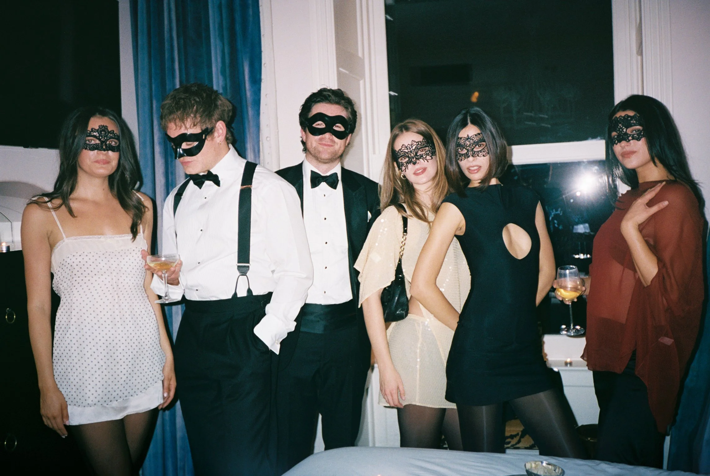 Group of six people dressed in formal attire, wearing black masks, standing close together at a party or celebration, holding drinks.