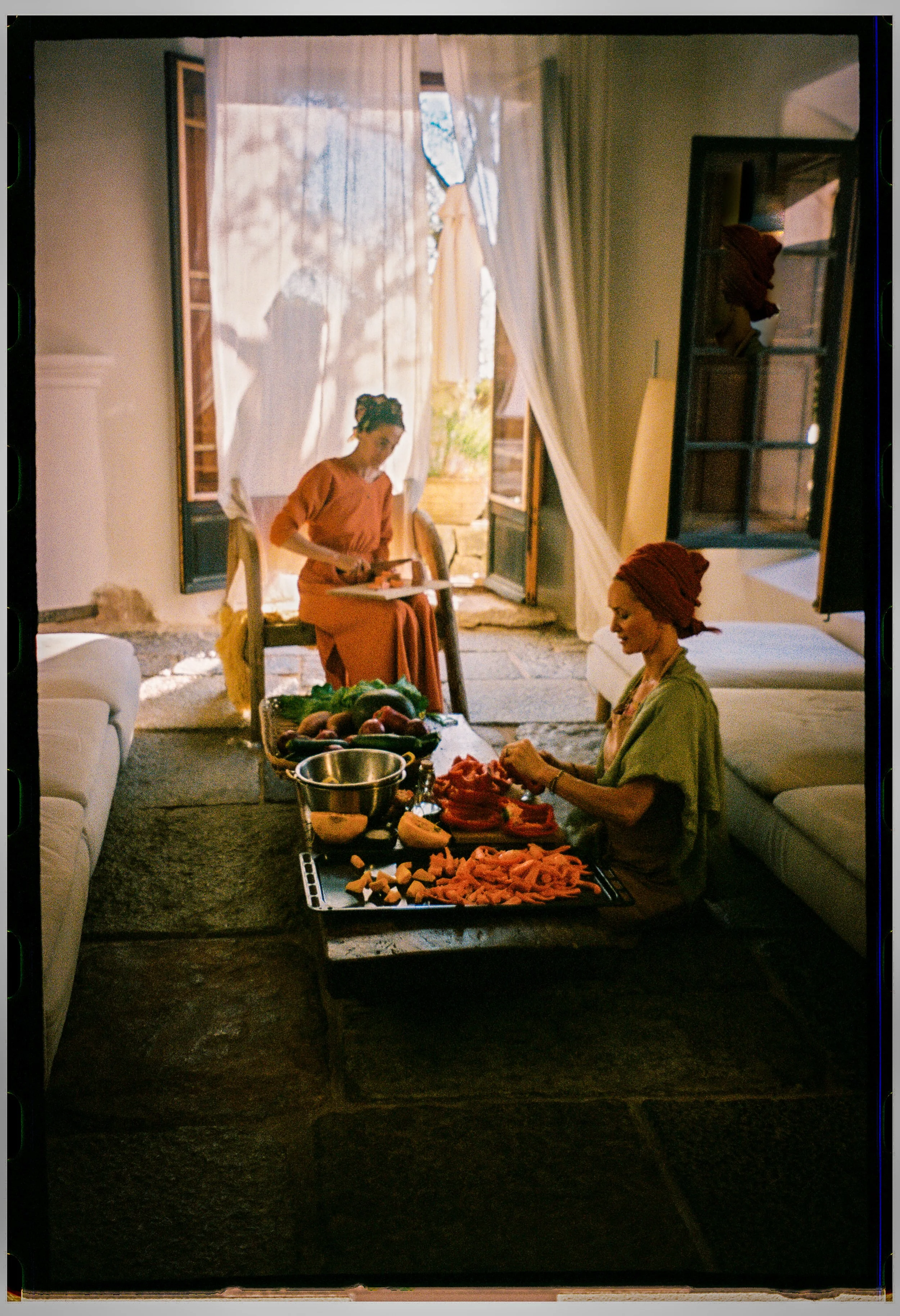 Two women preparing vegetables on a low table in a bright room with open doors and curtains, with a window showing trees outside.