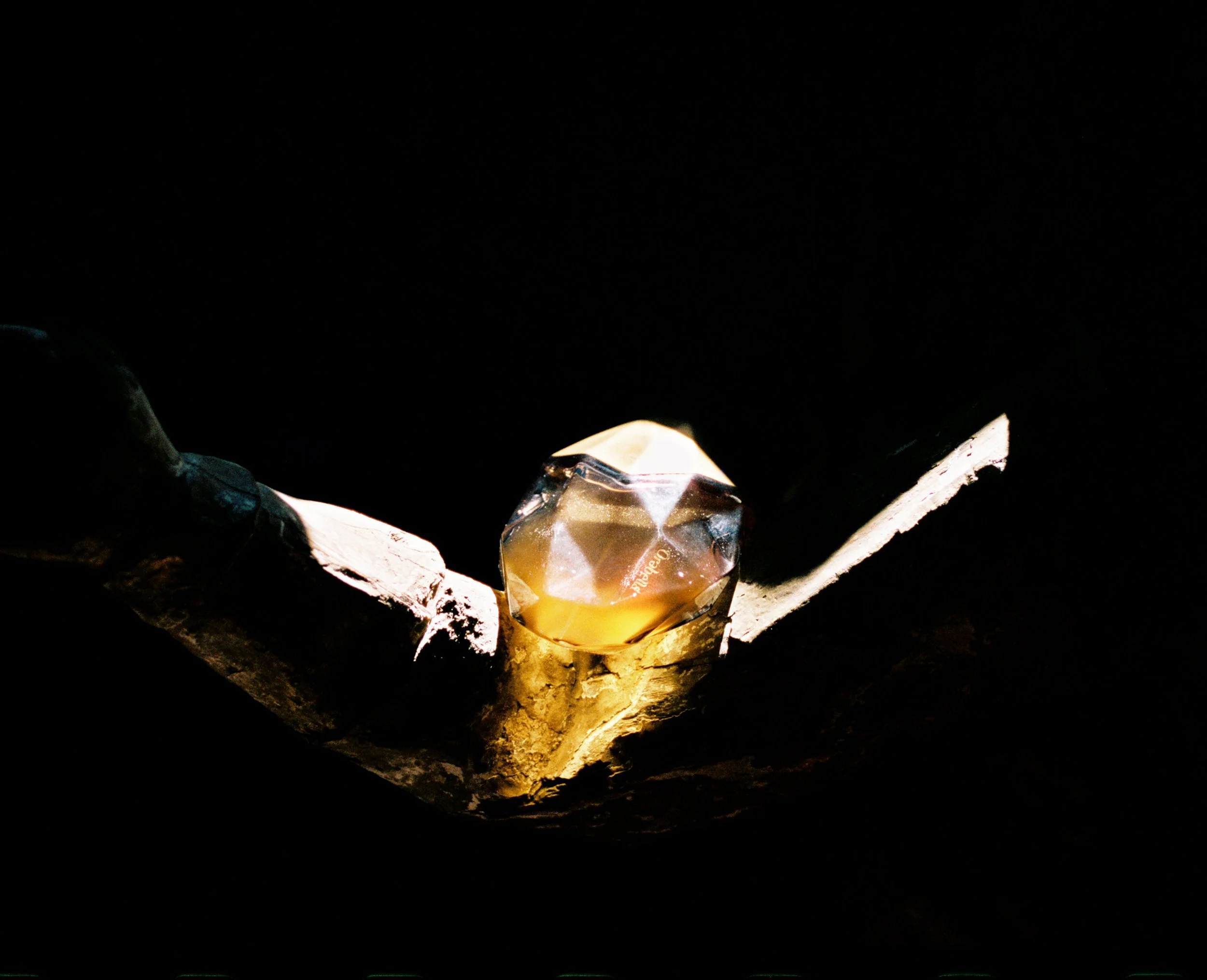 A metal object with a geometric, faceted surface resting on a piece of rough, burned wood inside a cast-iron skillet, with darkness surrounding them.