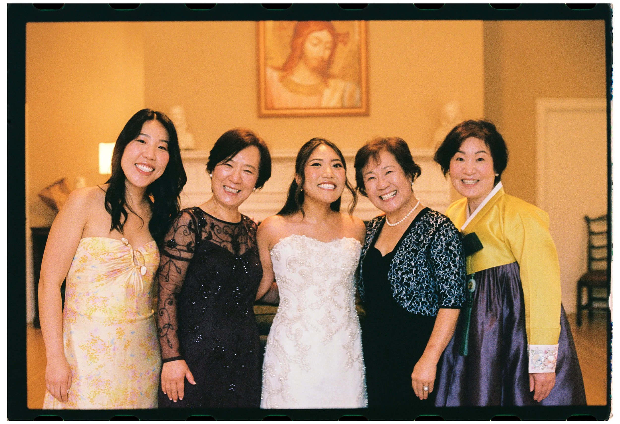 Five women smiling and standing together indoors, with a religious painting of Jesus Christ hanging on the wall behind them.