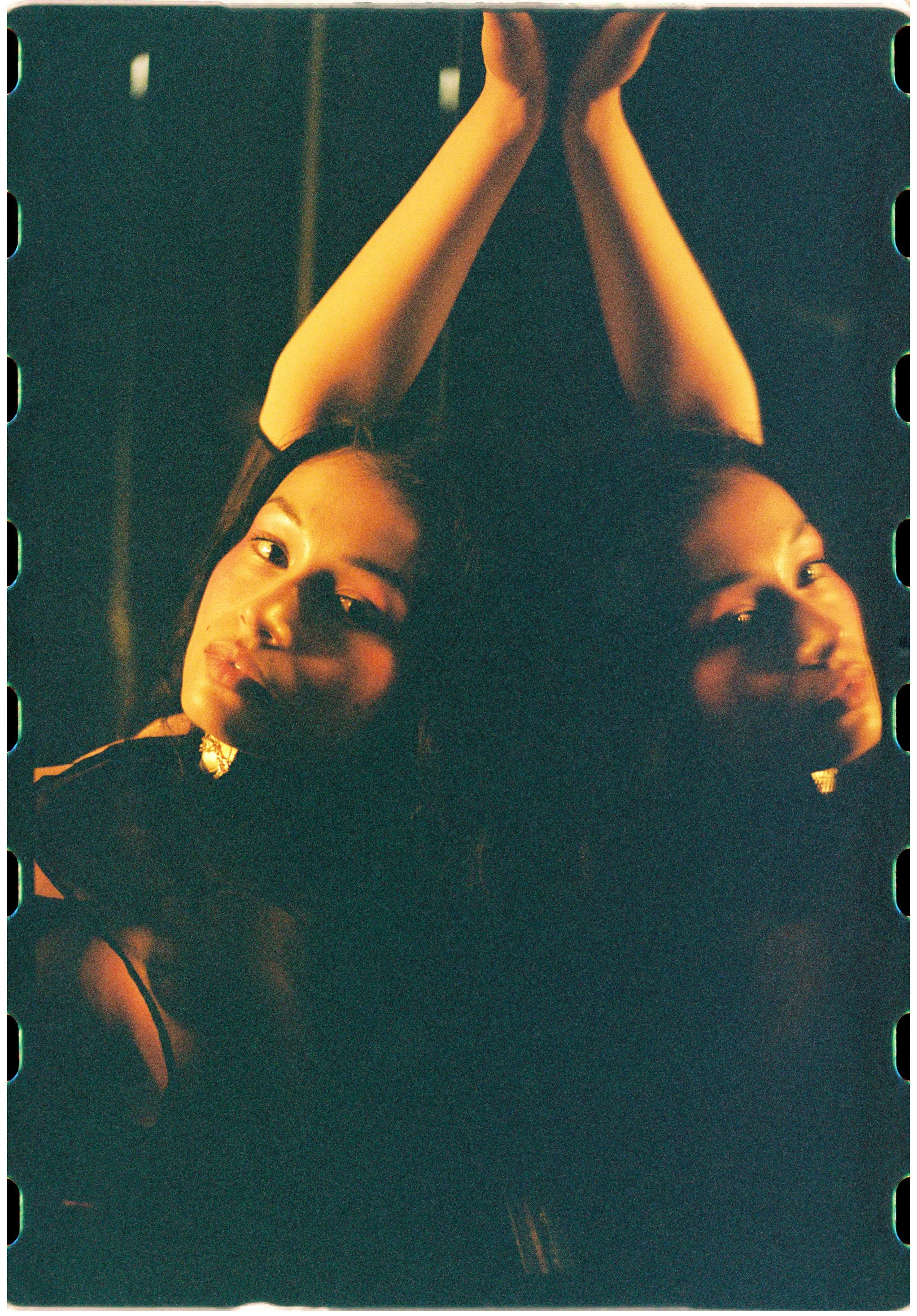 A woman with long dark hair is lying down with her arms stretched above her head, her hands resting on a white surface. She is looking directly at the camera with a neutral expression, and the lighting casts a warm glow on her face. The photo has a v
