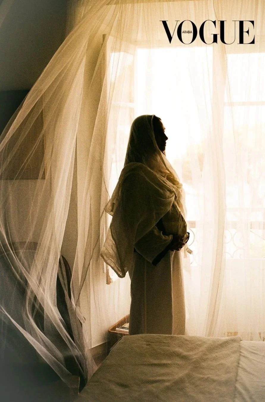 Silhouette of a woman standing beside a window with sheer curtains, backlit by sunlight, with "Vogue Arabia" text in the top right corner.