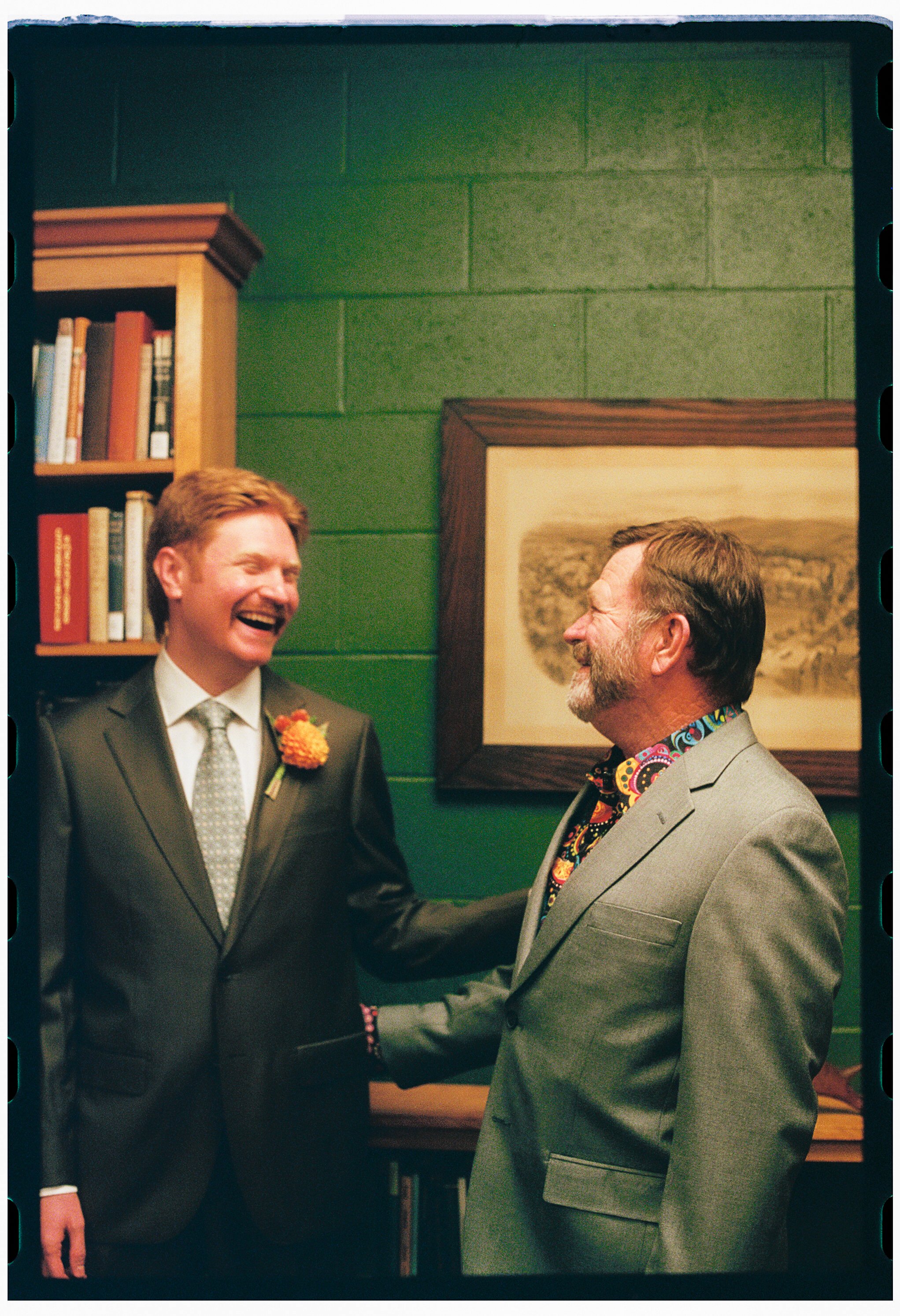 Two men in suits shaking hands and smiling at each other indoors, with a green wall and a framed picture in the background.