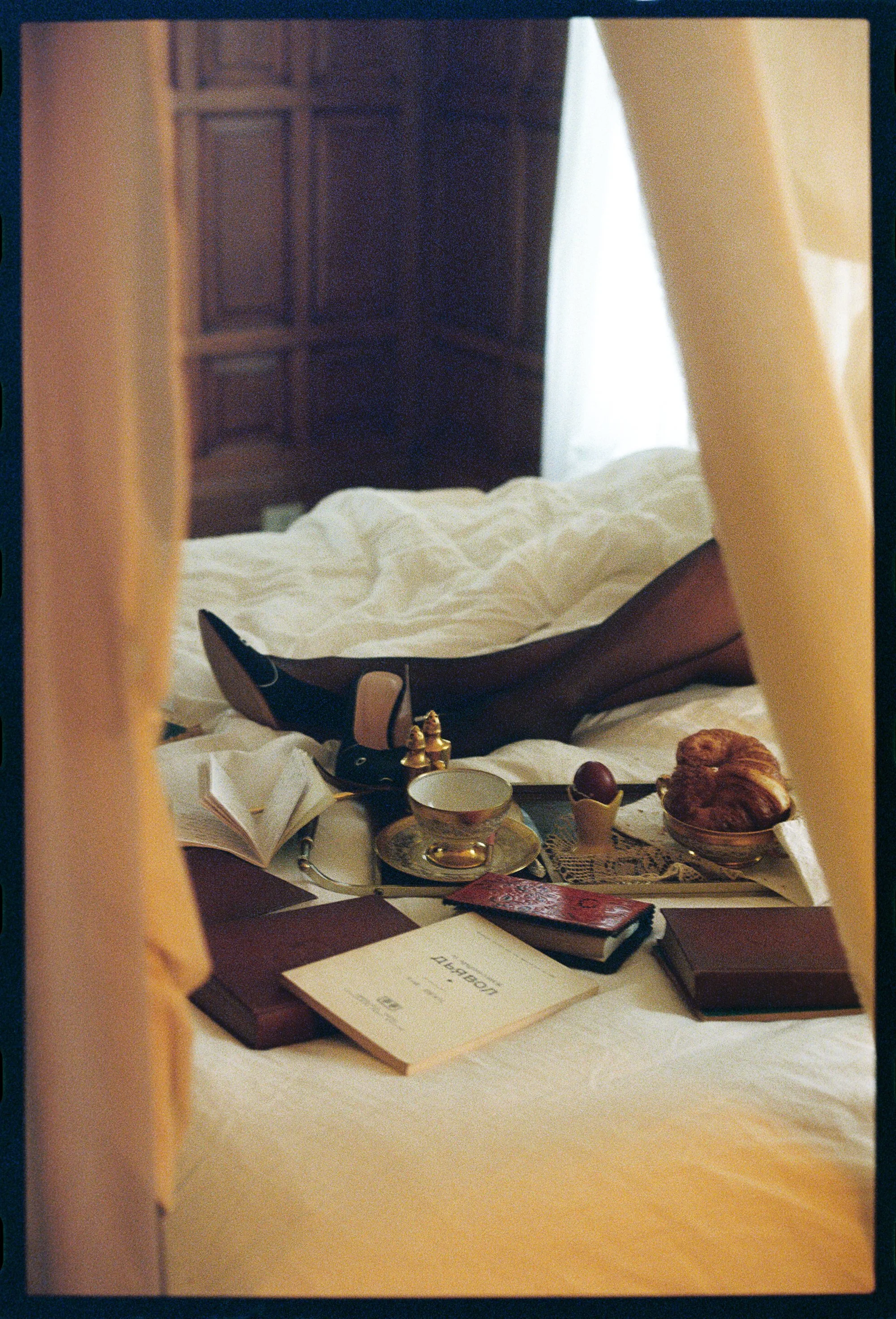 A breakfast tray on a bed with a croissant, a cup and saucer, a bowl of fruit, a notebook, a newspaper, and an open book, with a bedroom headboard in the background.