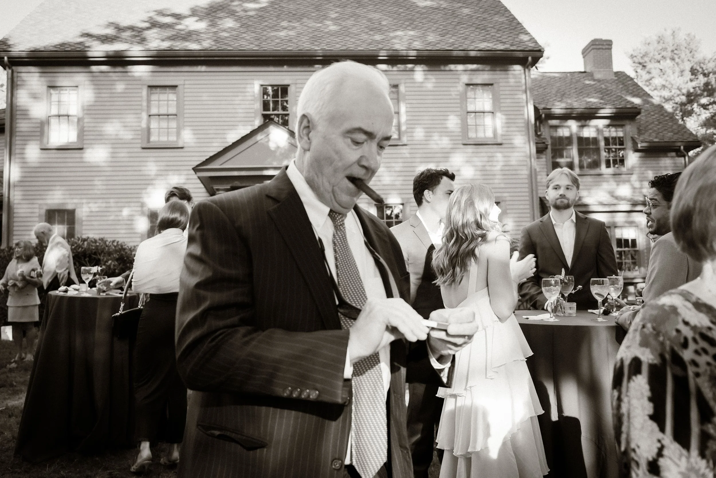 A classic retro black and white photo of an older man with a cigar in his mouth at an outdoor backyard wedding. Shot on 35mm film