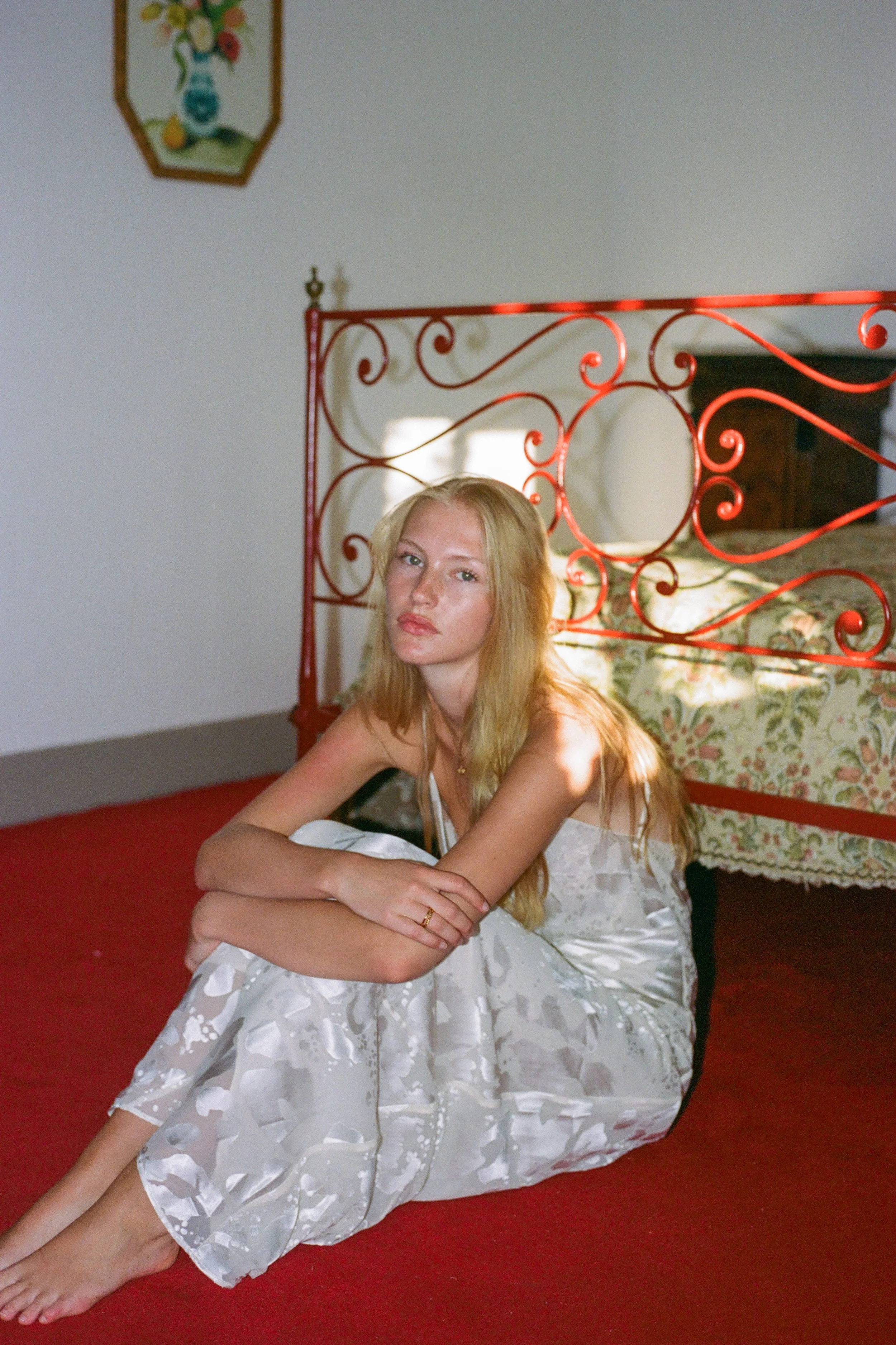 A young woman with long blonde hair wearing a white dress with floral patterns, sitting on the floor and leaning on her arm in a bedroom with a red metal headboard, floral bedding, and a picture of a vase with flowers on the wall.