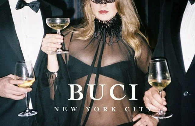A woman in a sheer black top with a lace collar holding a glass of champagne at a fancy event. Two men in tuxedos are also holding glasses of champagne on either side of her. The words 'BUCI New York City' are overlaid on the image.