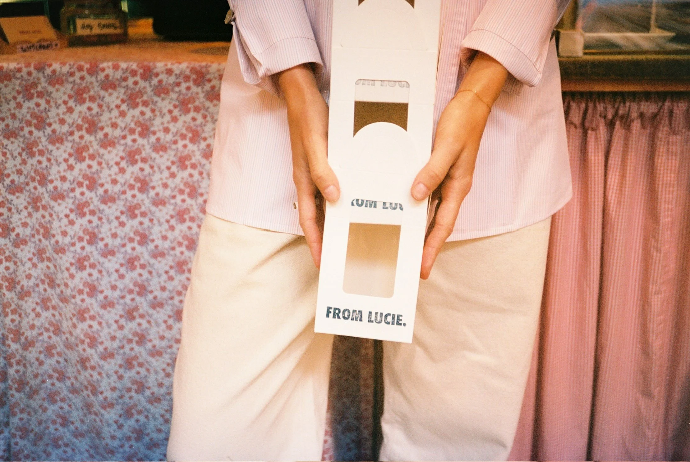 A person in a pink and white striped shirt is holding a white cardboard sign that says "FROM LUCIE." The background features a table draped with a pink and red floral patterned cloth, and books and other items are visible. The person's face is not sh
