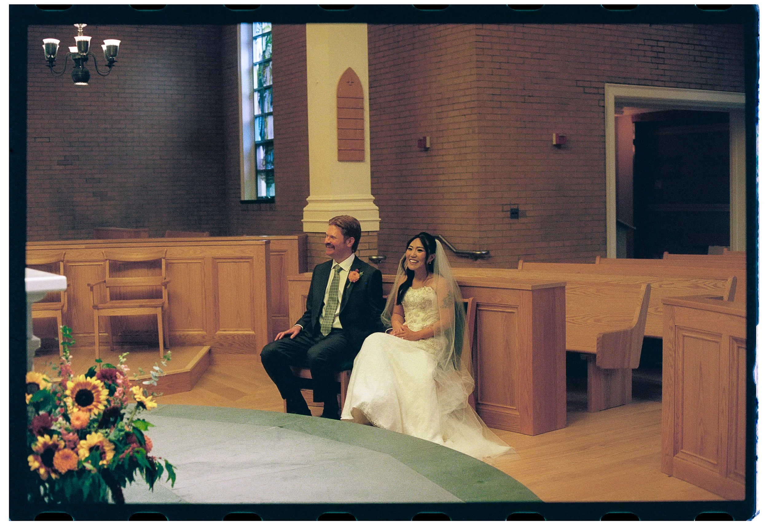 A bride and groom sitting in a church pew during a wedding ceremony, smiling, with brick walls and wooden pews in the background, and flowers in the foreground.