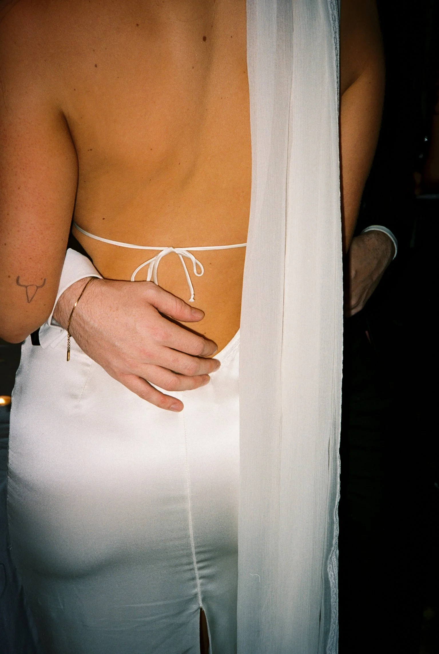 Close-up of a woman's back and side, dressed in a white top with a tie string and white pants, with a tattoo of a bull's head on her arm