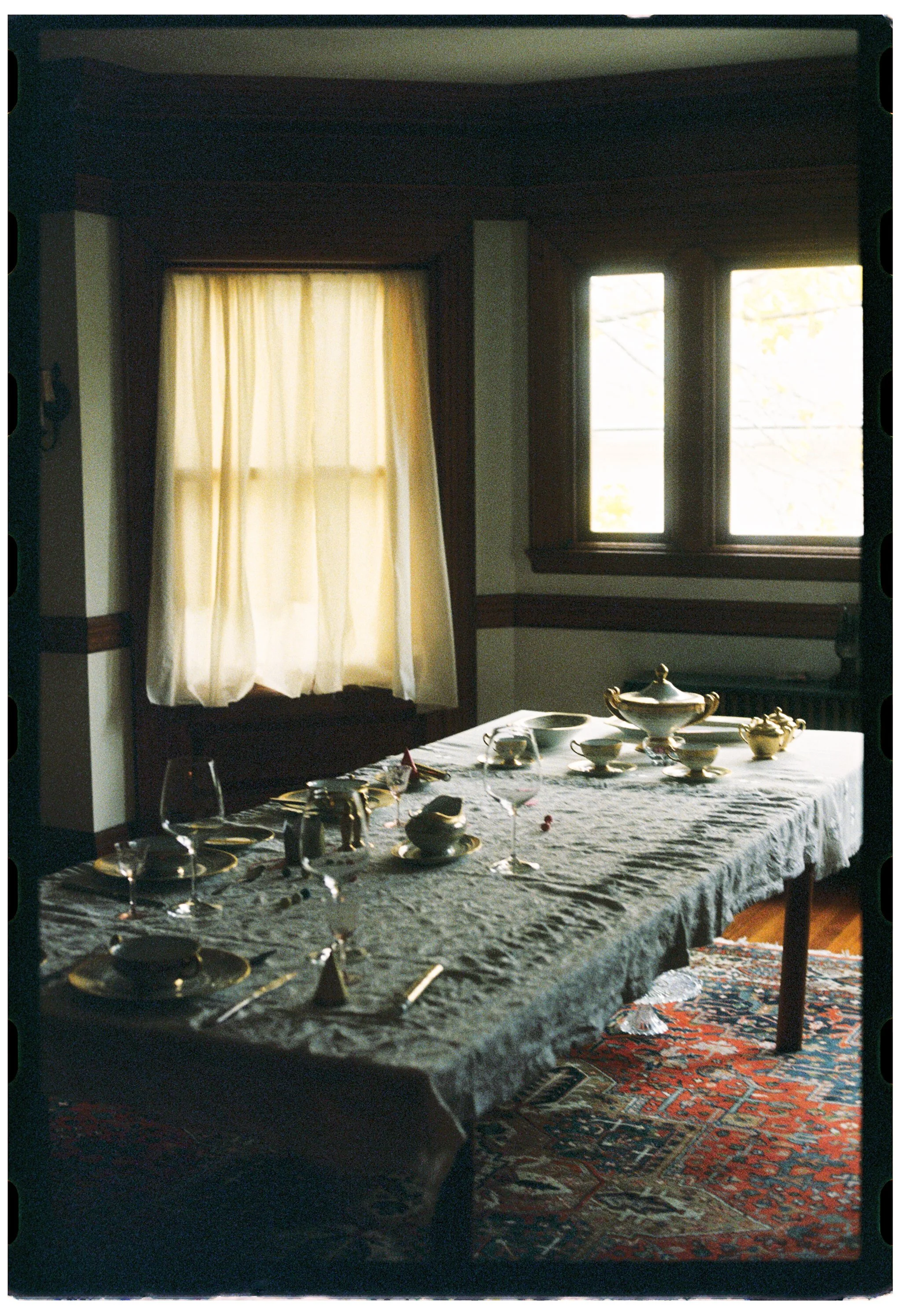 A dining room with a table set with fine china, glassware, and silverware, covered with a lace tablecloth. Large windows with curtains let in natural light, and a patterned rug covers the wooden floor.