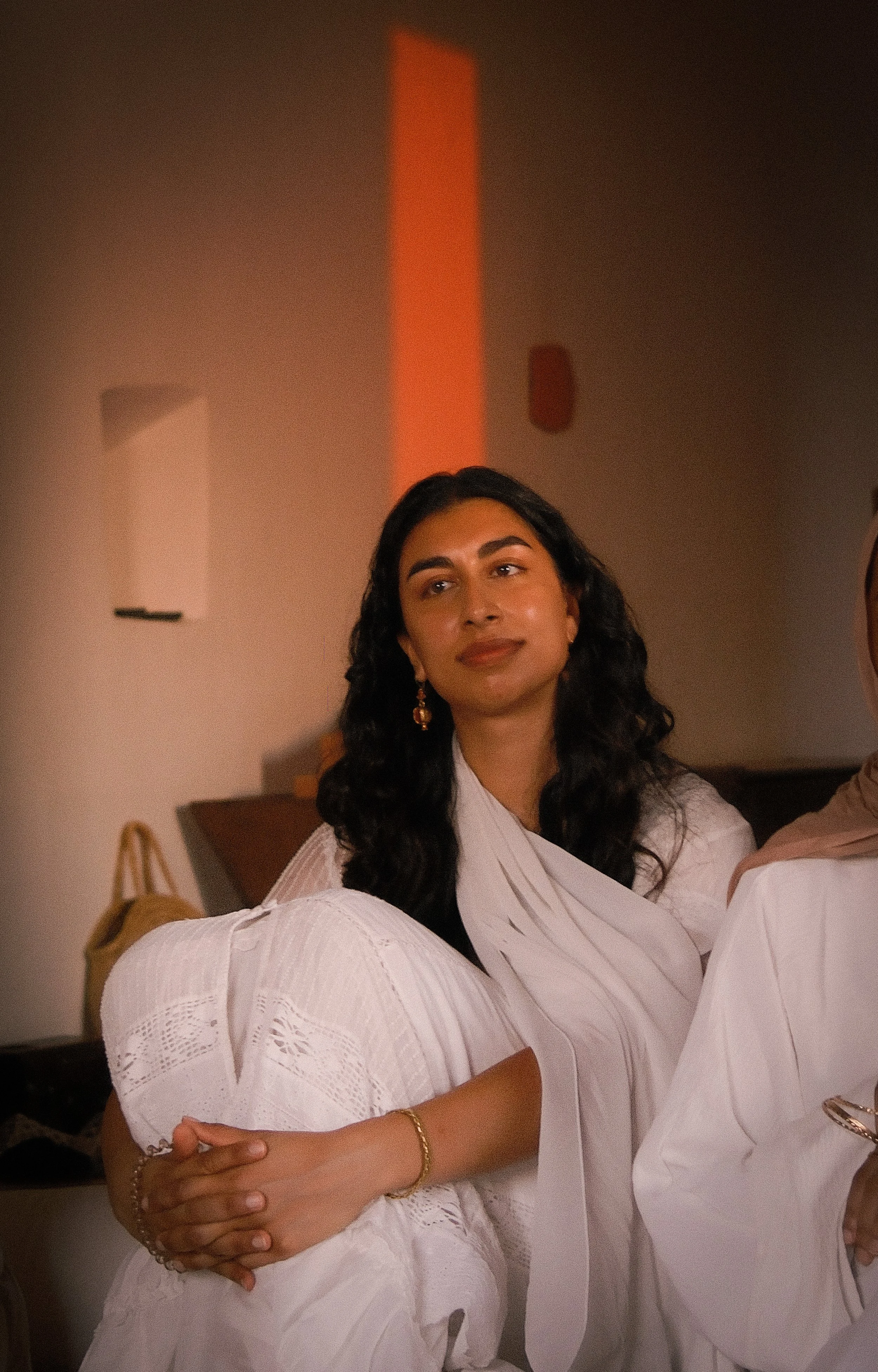 A woman with dark, curly hair and earrings, dressed in traditional light-colored attire, standing indoors with a gentle smile. She is embracing someone wearing a similar outfit.