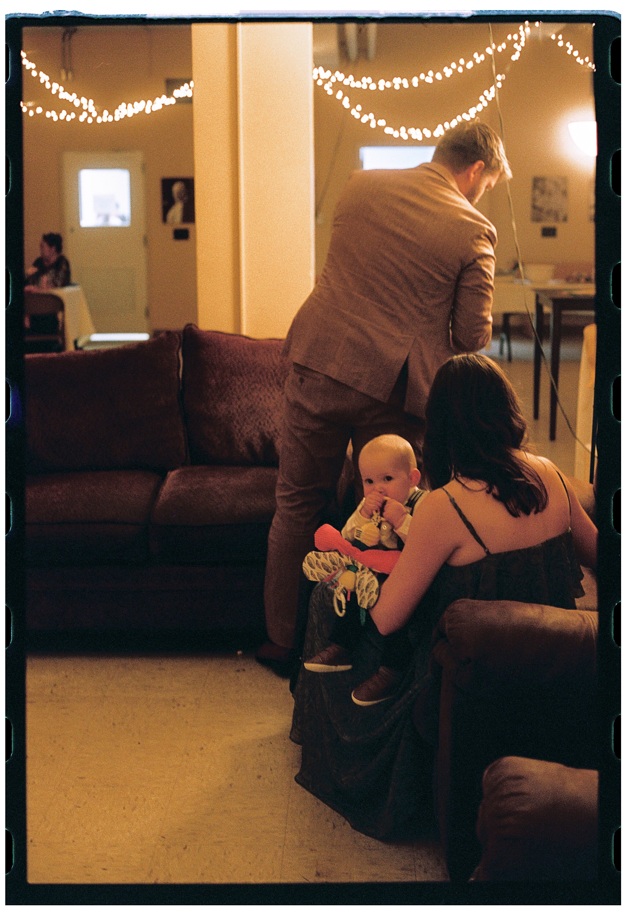 A woman with dark hair holding a baby with a pacifier, a man in a brown blazer standing near them, in a room decorated with string lights, and a woman sitting at a table in the background.
