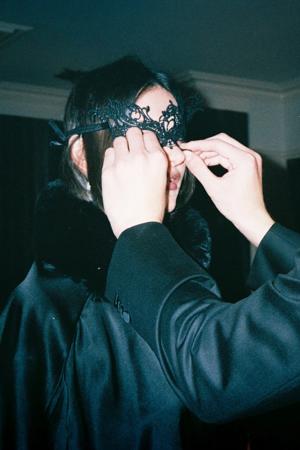Person putting on a black lace mask while someone helps adjust it, indoors with dark surroundings.