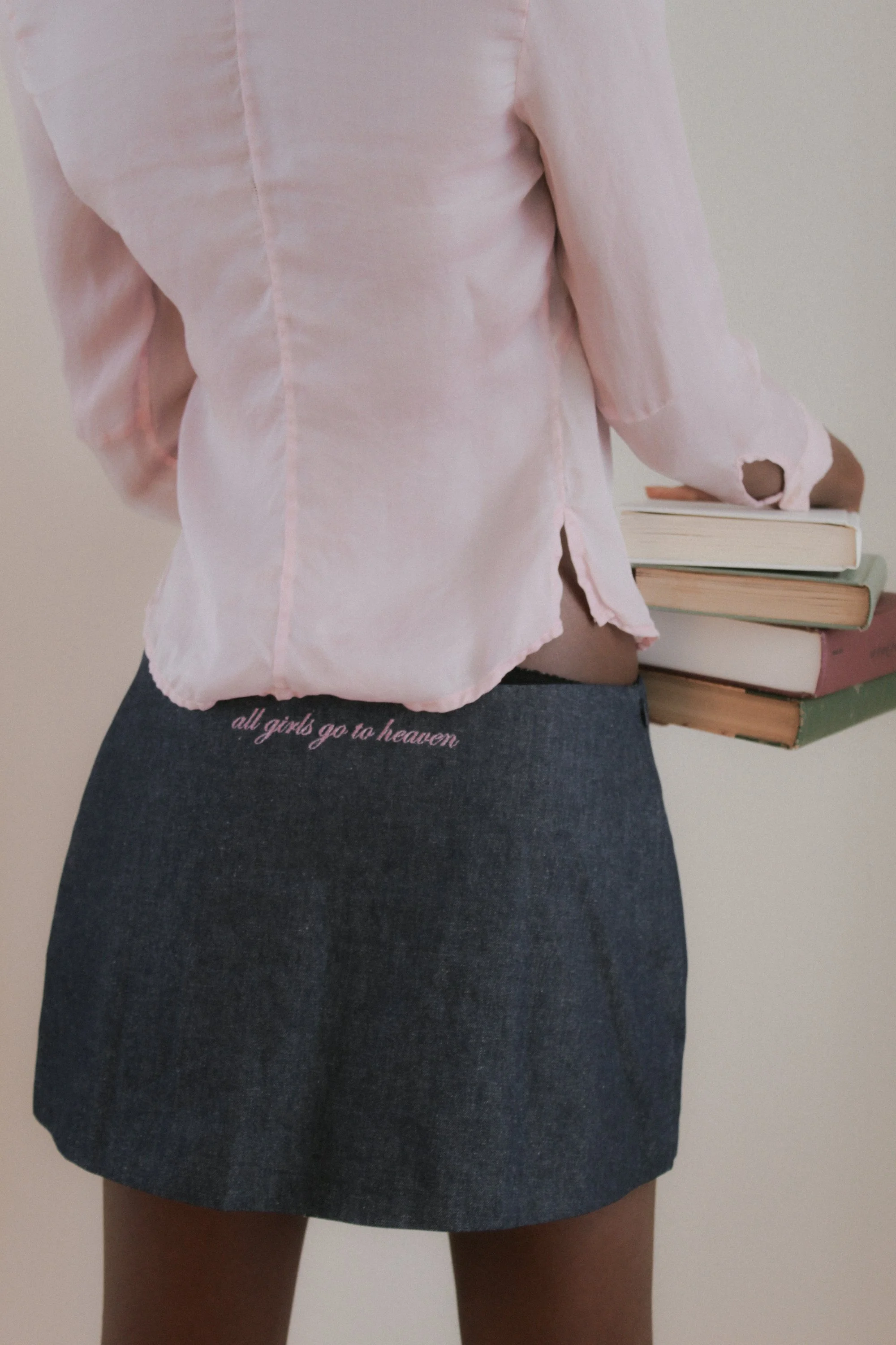 A person wearing a light pink long-sleeve shirt and a dark denim skirt, holding a stack of five books. The skirt has the phrase 'all girls go to heaven' embroidered near the waistline.