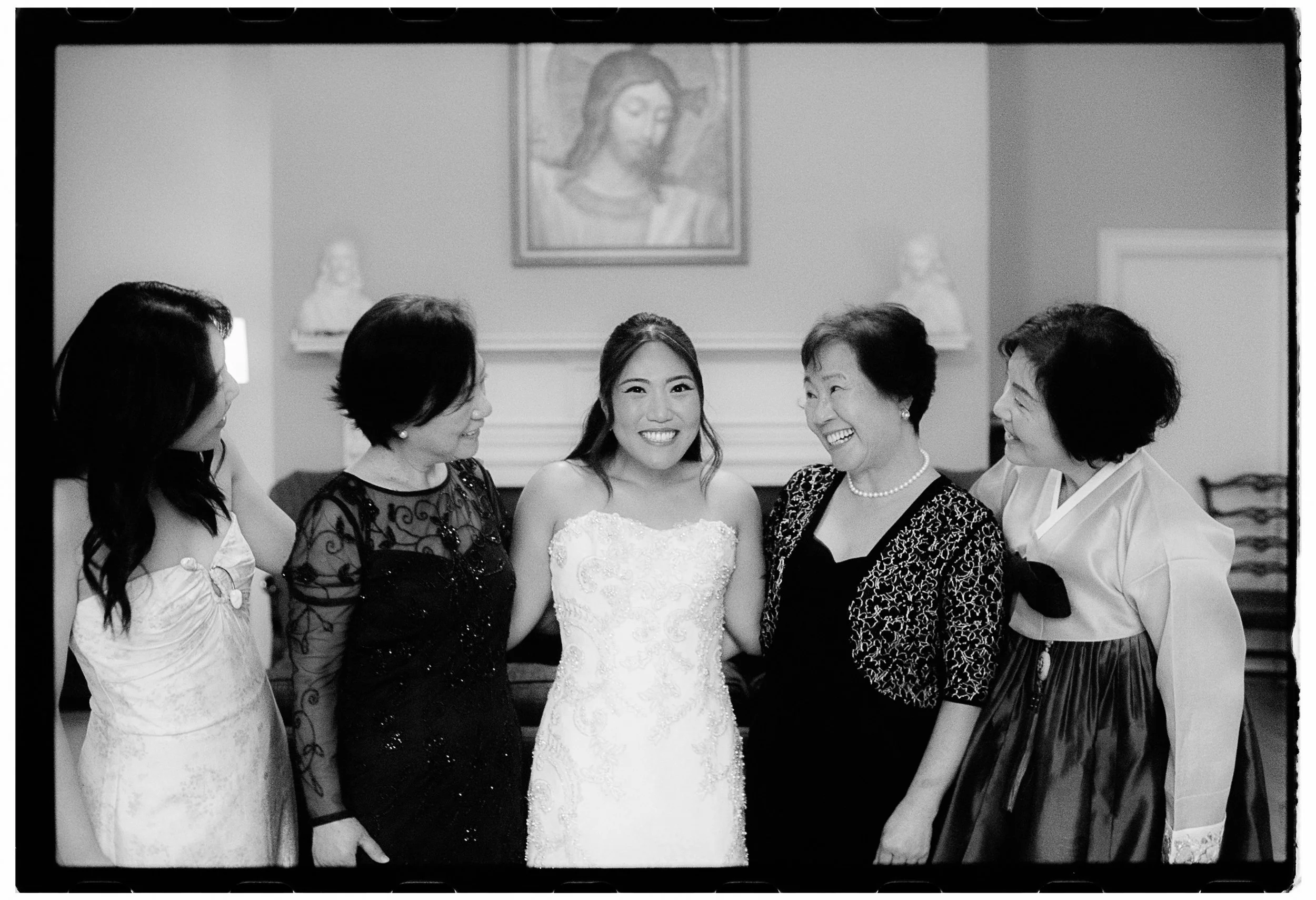 Black and white photo of six women, with the woman in the middle wearing a wedding dress, surrounded by five women in formal dresses, in a room with a painting of Jesus Christ on the wall behind them.
