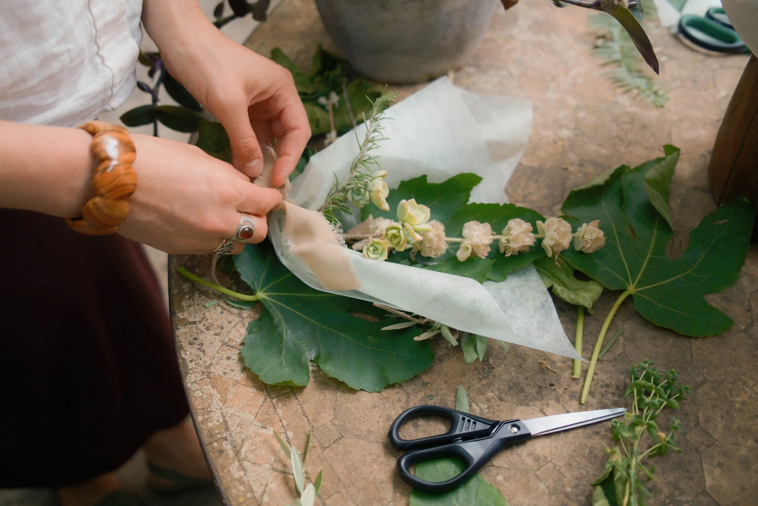 Person working on floral arrangement with flowers, leaves, scissors, and wrapping paper on a kitchen counter.