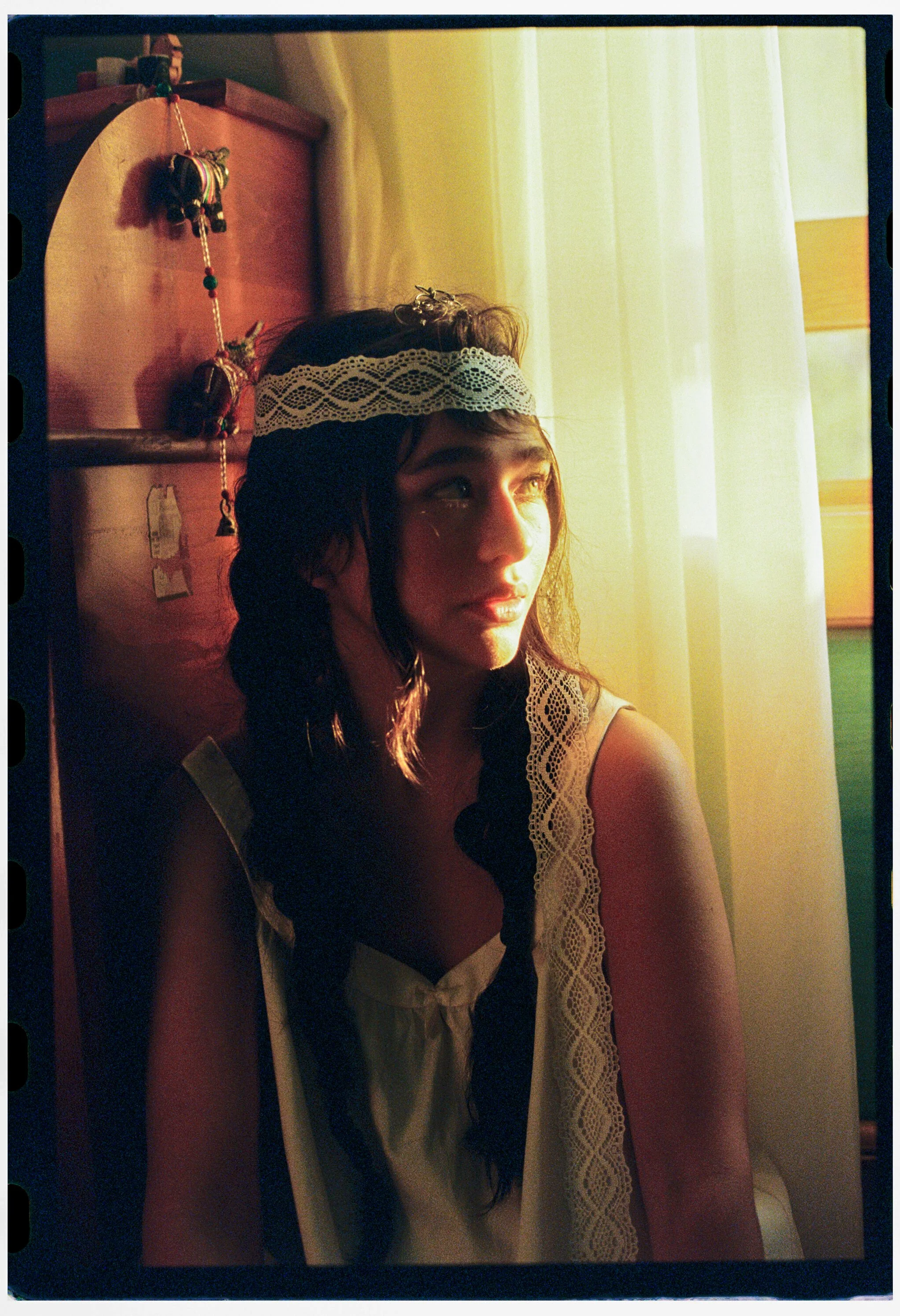 A young woman with dark hair, wearing a lace headband and a sleeveless dress with lace details, is looking out a window as sunlight illuminates her face.