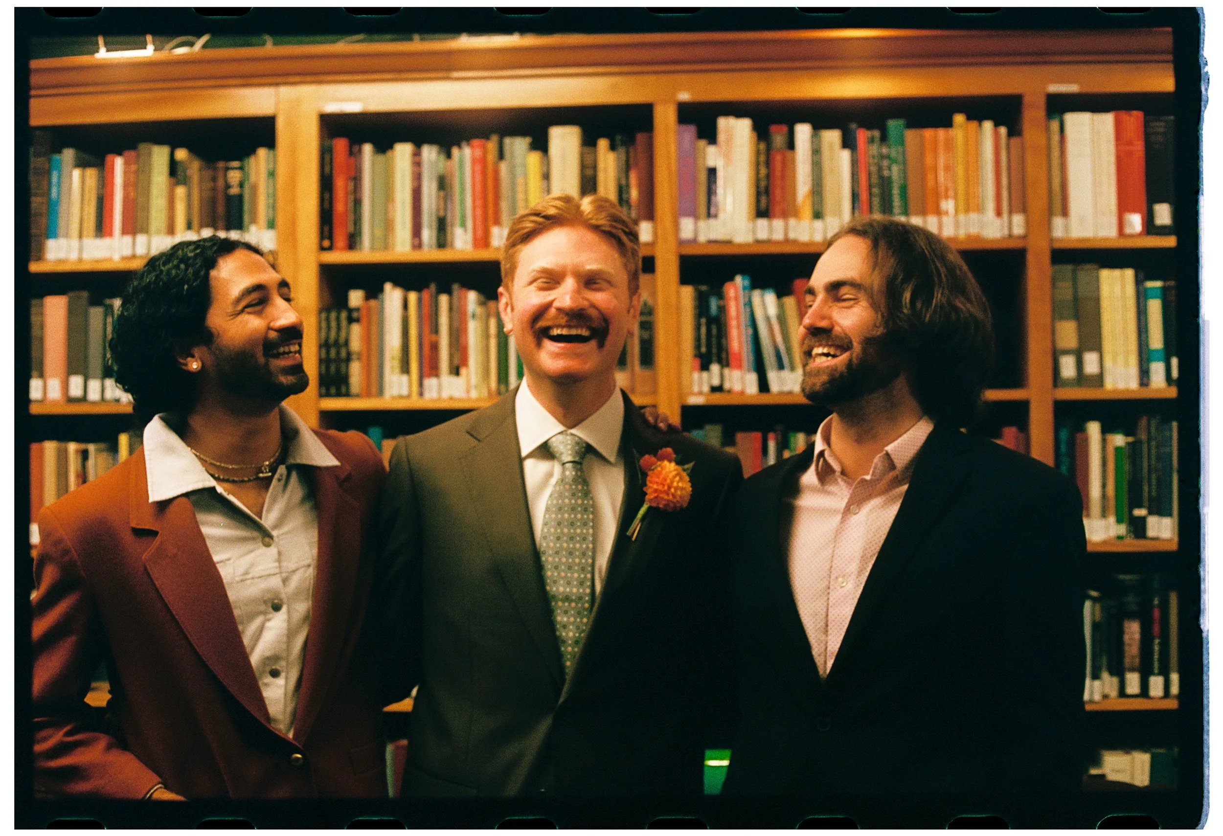 Three people smiling and laughing together in front of a bookshelf filled with books, in an indoor setting.