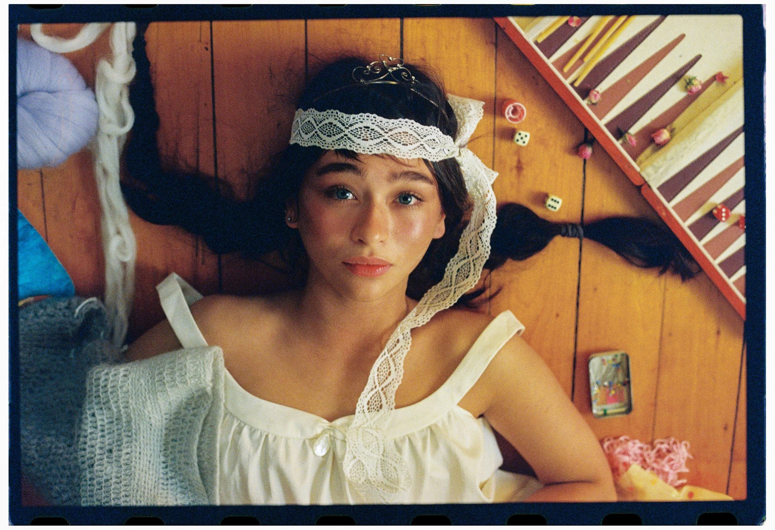 A young woman with blue eyes and long dark hair, lying on a wooden floor. She is wearing a white top with lace details, a lace headband, and a decorative hair accessory. Surrounding her are items including yarn, dice, a small Lego piece, a smartphone