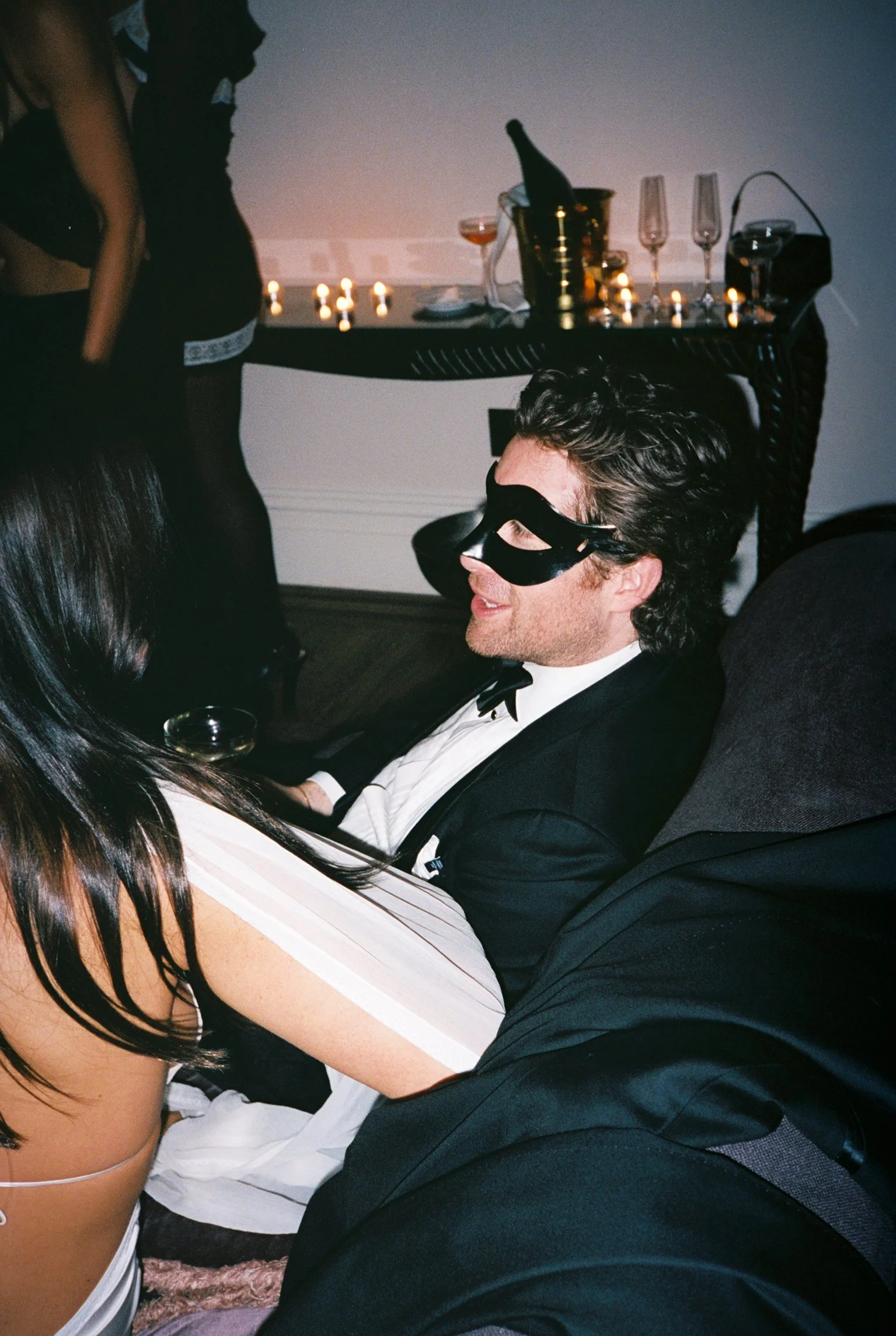 A man in a tuxedo and sleep mask sitting on the floor at a party, with a woman in lingerie lying next to him. There is a table with a bottle of champagne and glasses behind them, and candles lit on the table.