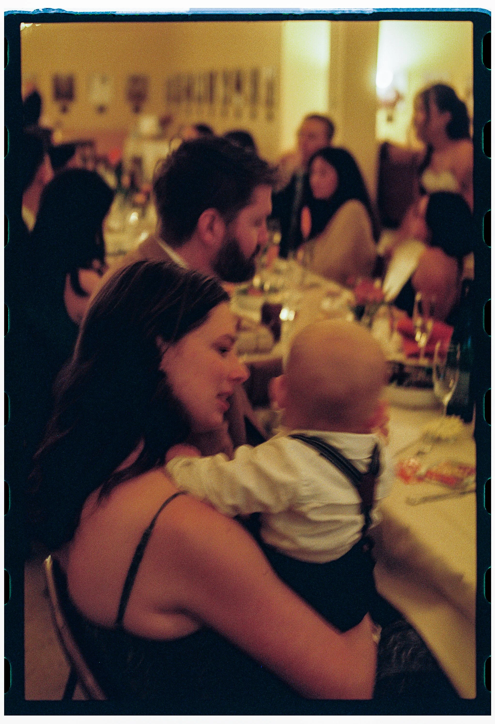 People sitting at a dinner table in a warmly lit room, engaged in conversation and socializing, with some individuals smiling and others listening attentively.