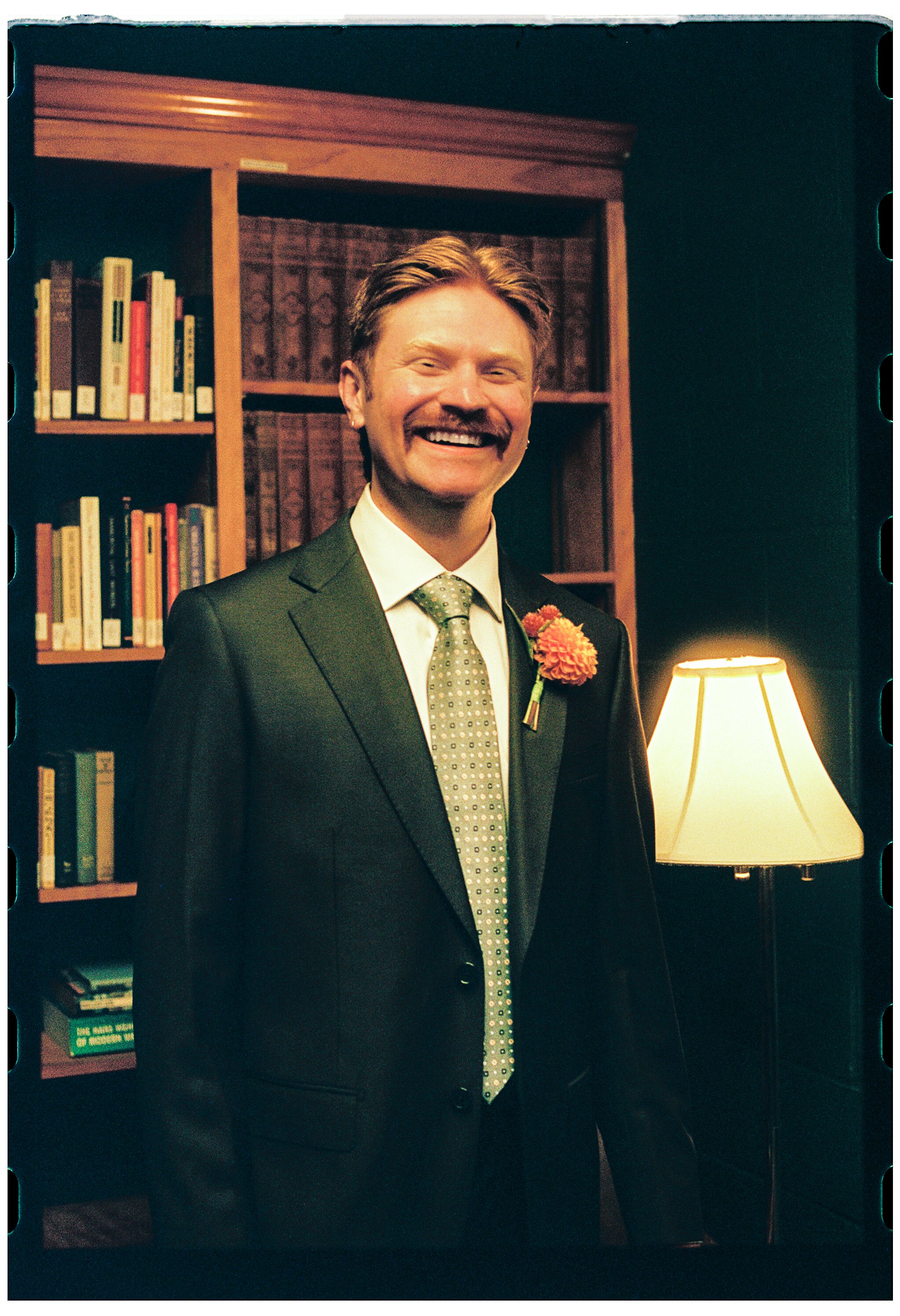 A smiling man in a suit standing indoors near a bookshelf and a table lamp.
