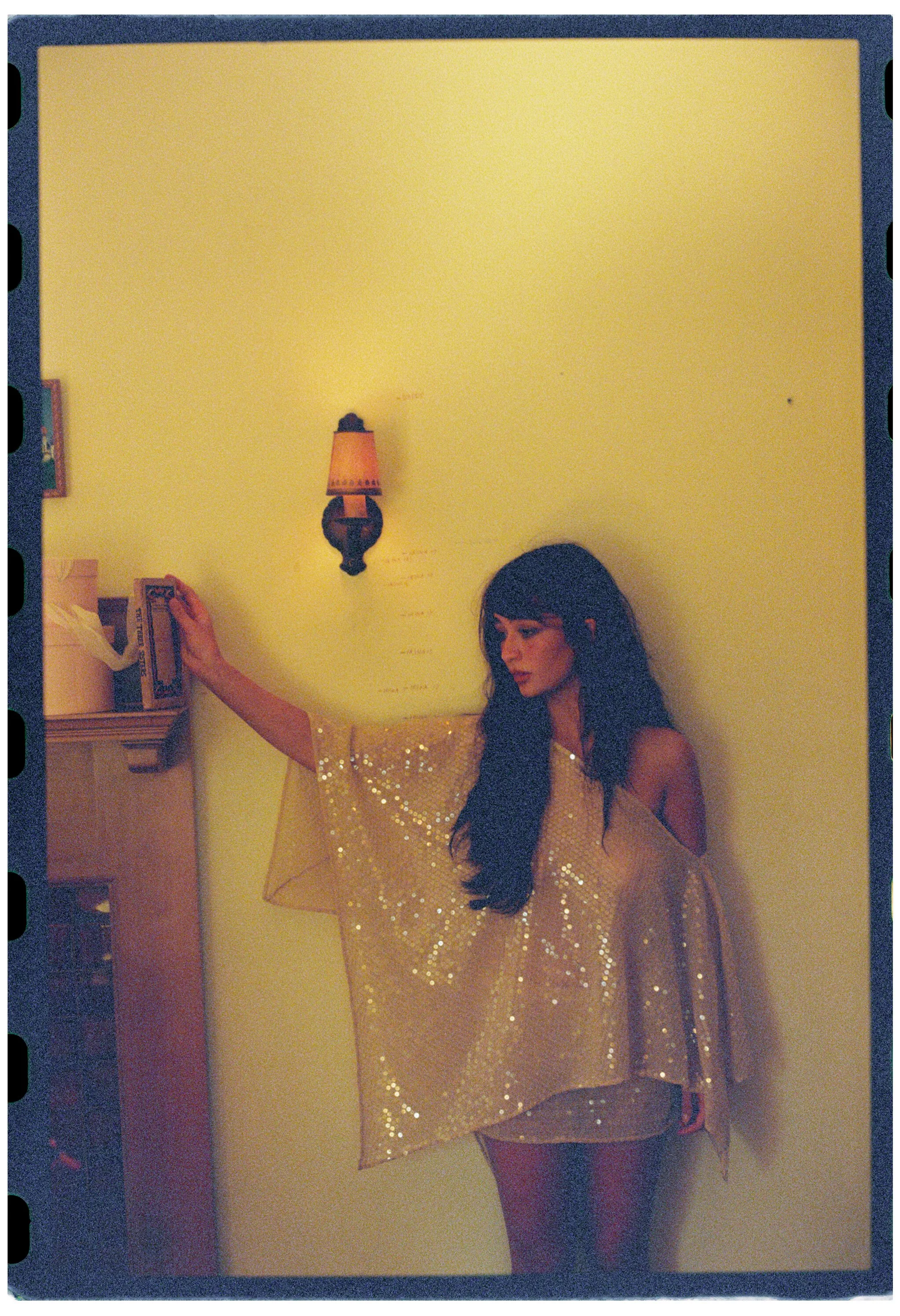 A woman with long dark hair in a shiny gold dress with sheer sleeves, standing against a yellow wall with a small wall lamp.