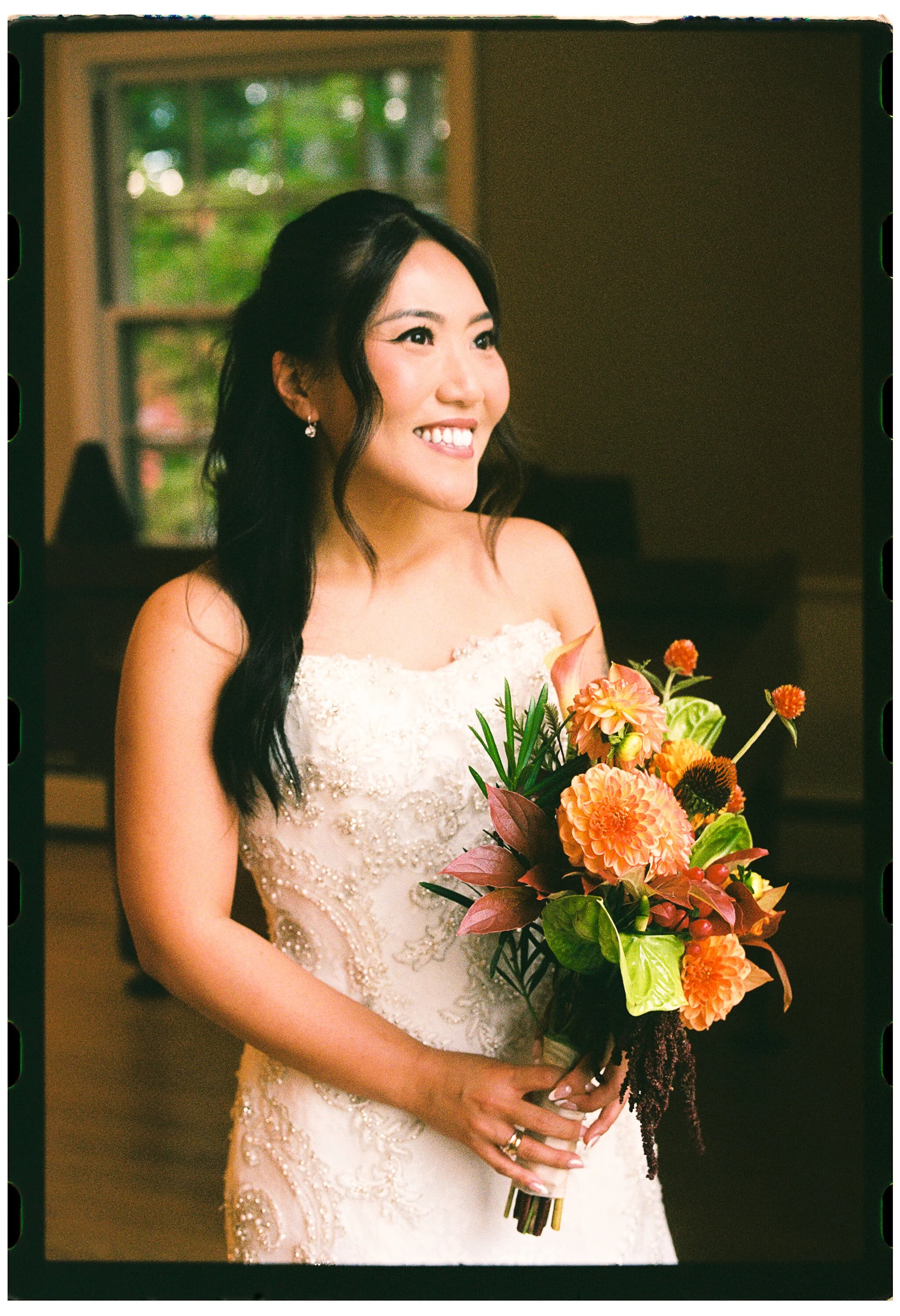 A smiling woman in a wedding dress holding a colorful bouquet of flowers indoors with a window in the background.