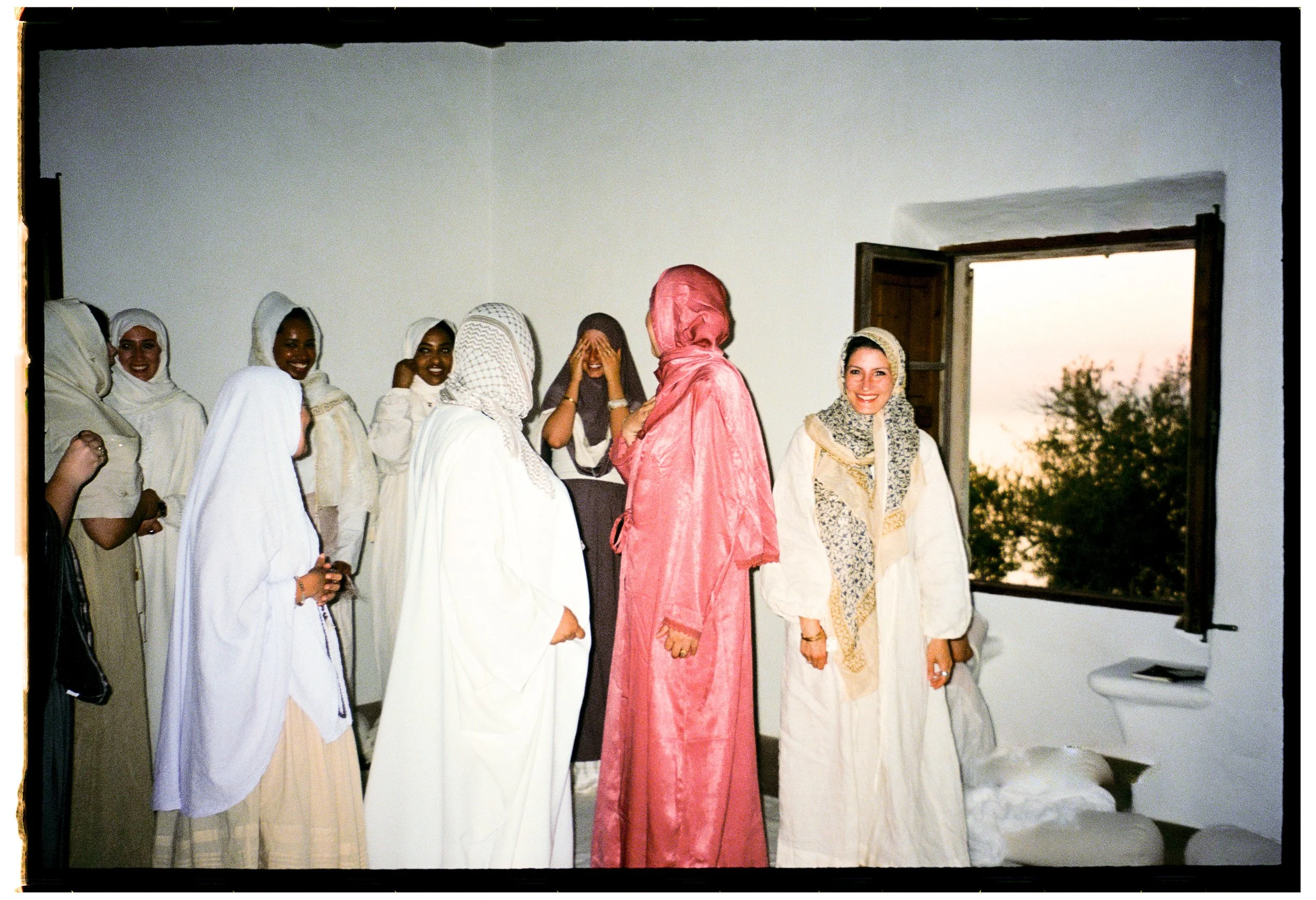 A group of women in traditional and modest clothing gathered in a room, some wearing hijabs, with a window open showing trees and an evening sky in the background.