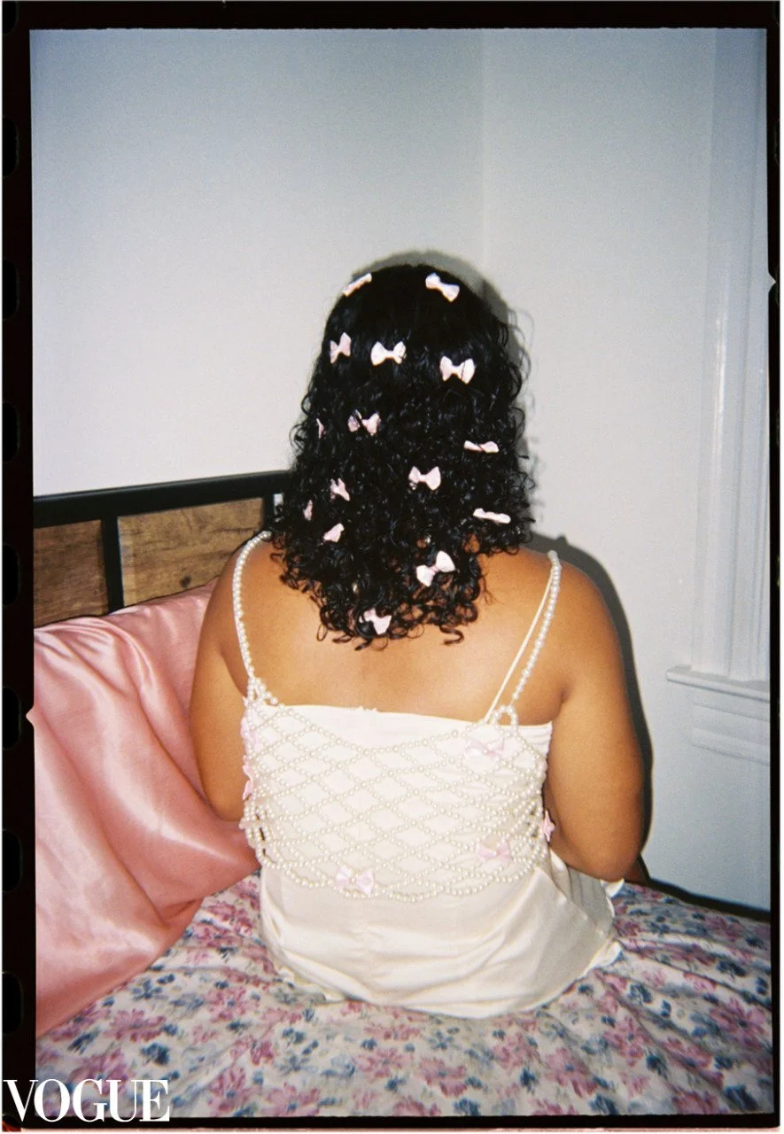 Woman with curly hair adorned with pink and white bows, sitting on a bed with pink satin pillow and floral bedding, wearing a cream-colored nightdress with pearl embellishments, facing away from the camera.
