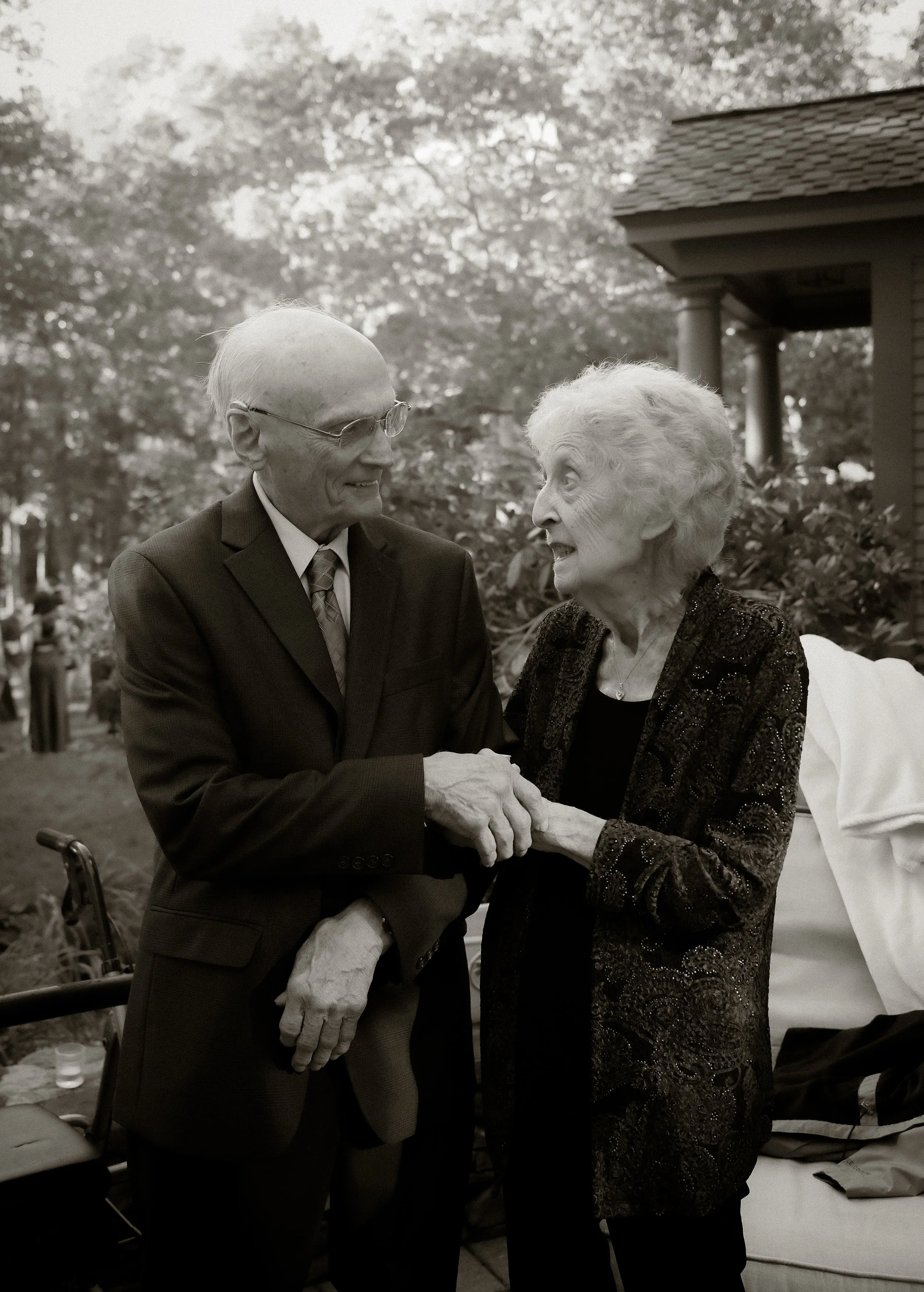 An elderly man and woman, grandparents of the bride, hold hands and smile lovingly at each other outdoors at a large backyard wedding in massachusetts. Shot on 35mm film