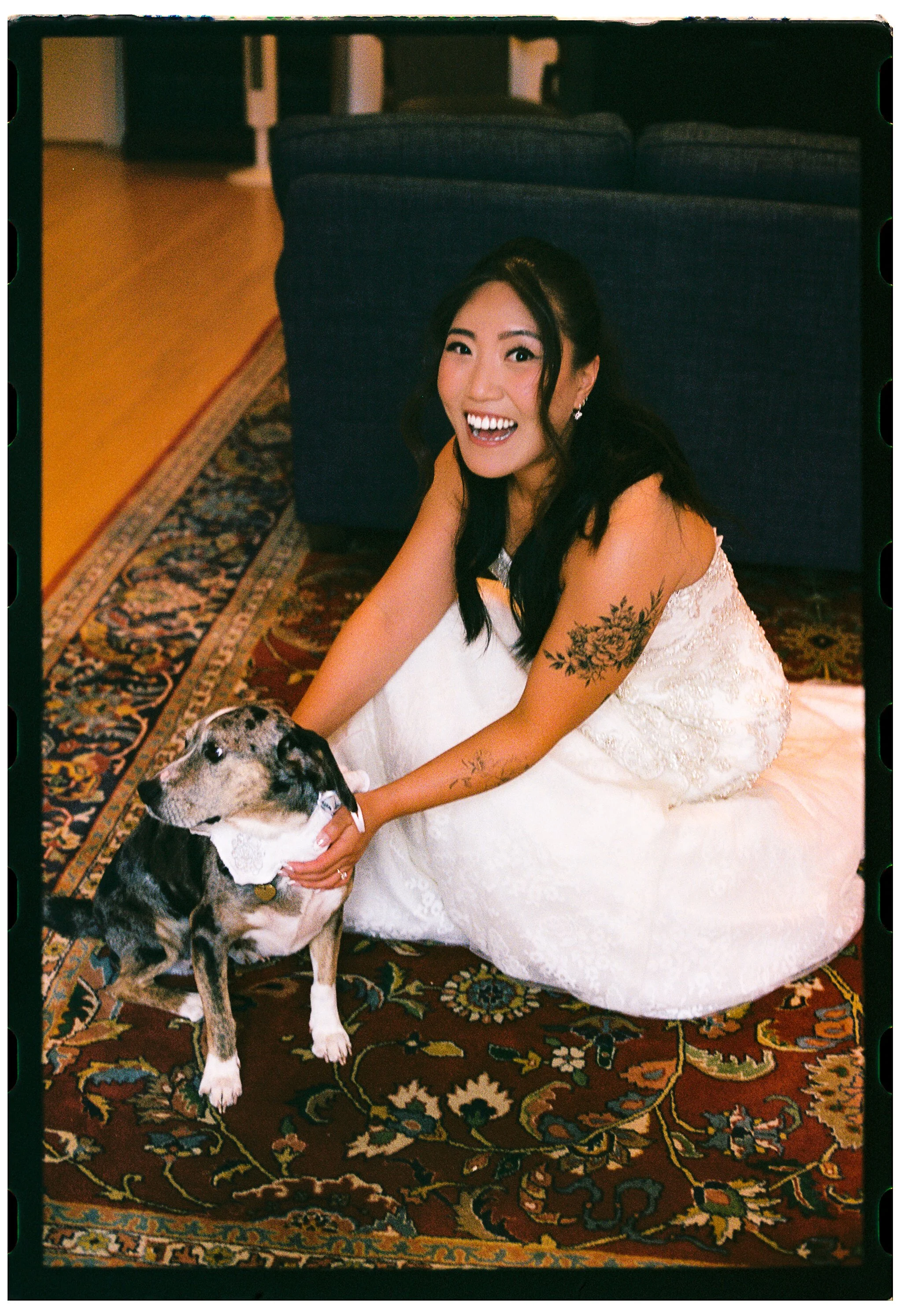 A woman in a white dress, smiling, sitting on a colorful patterned rug indoors, with a dog in front of her wearing a white lace collar.