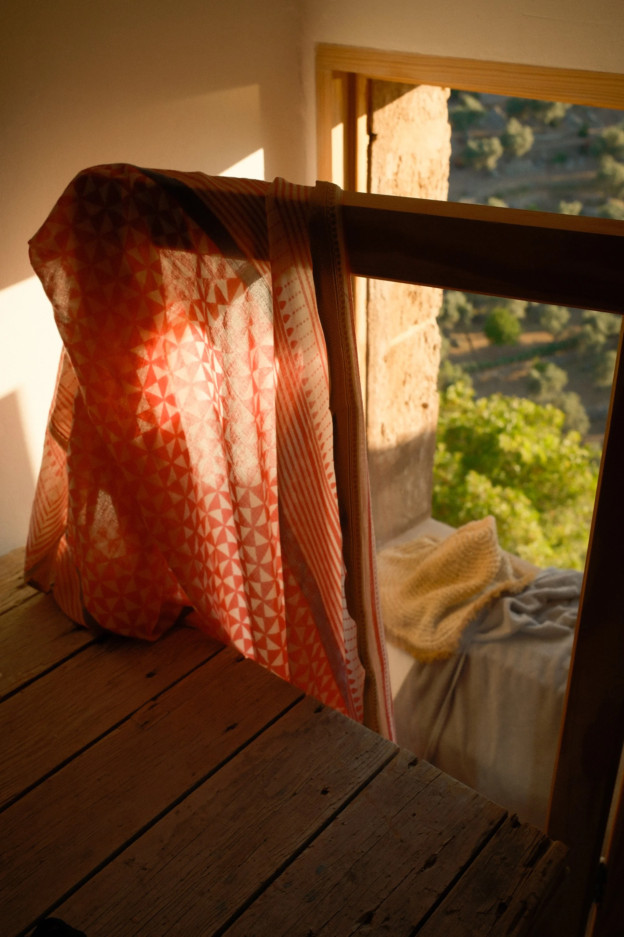 A mirror reflecting a window with a view of trees and a landscape outside, a wooden table, a blanket, and a curtain with geometric patterns.