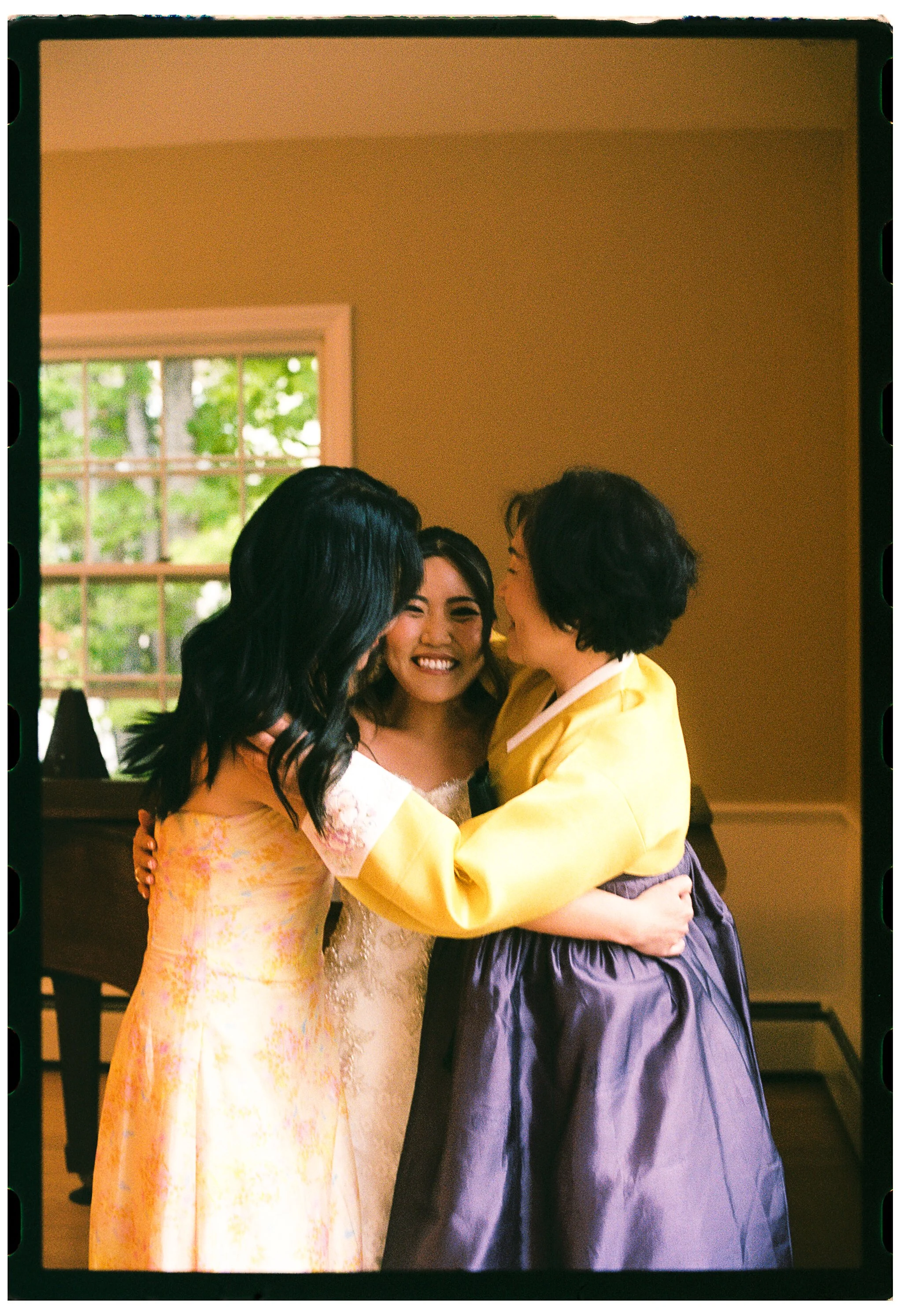 Three women embrace and smile warmly in a room with a window in the background.