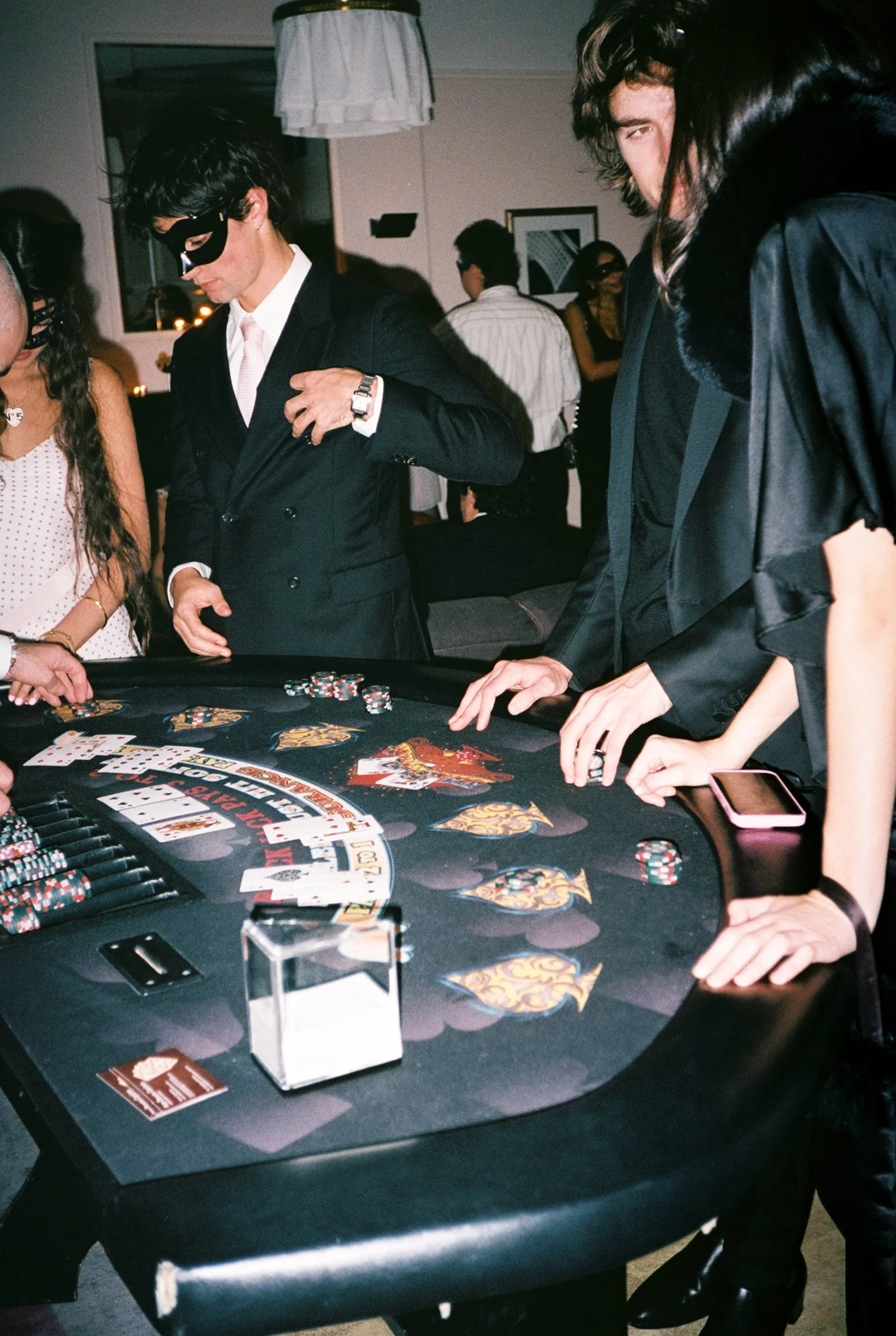 People playing blackjack at a casino table; some are dressed in formal attire including suits, one person is wearing a mask, and there are chips, cards, and a payout slip on the table.